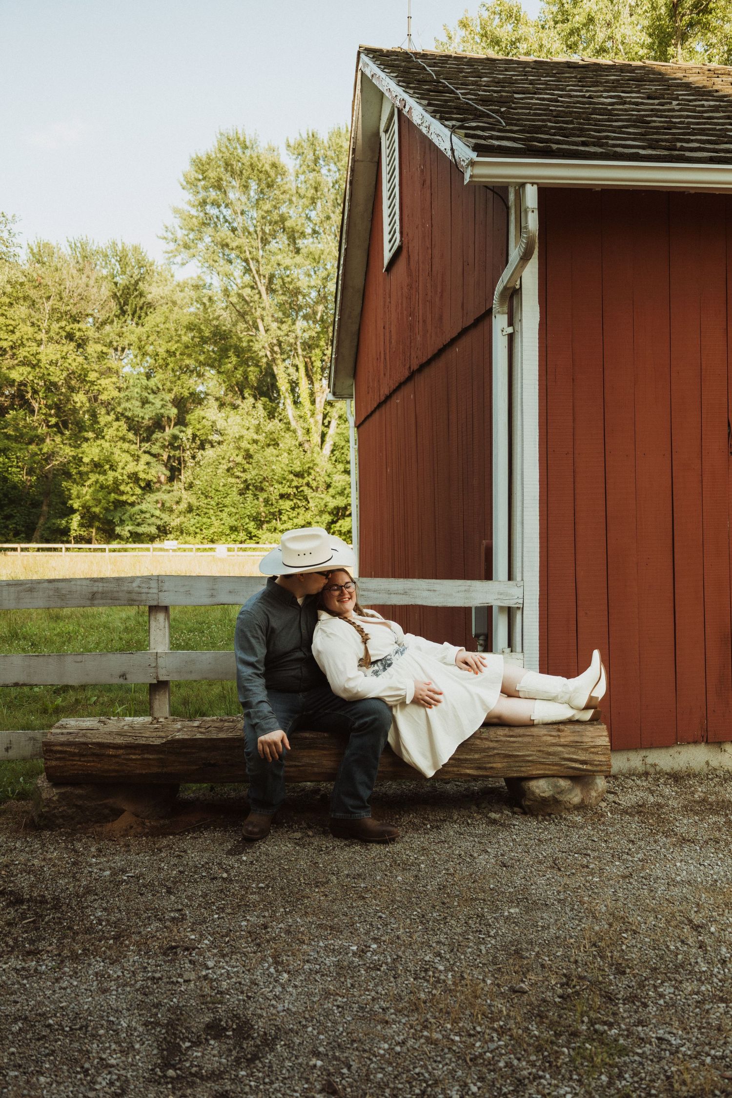 A couple shares a tender moment while resting on a wooden bench outside a rustic red barn on a sunny day.