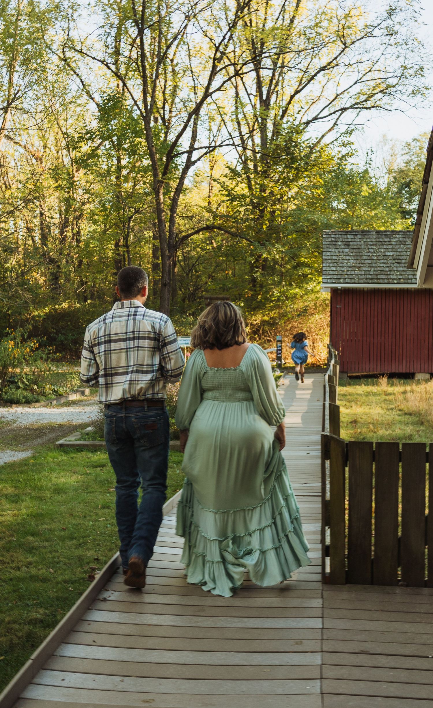 People walking on a wooden boardwalk path through autumn trees near a red barn building on a sunny day.