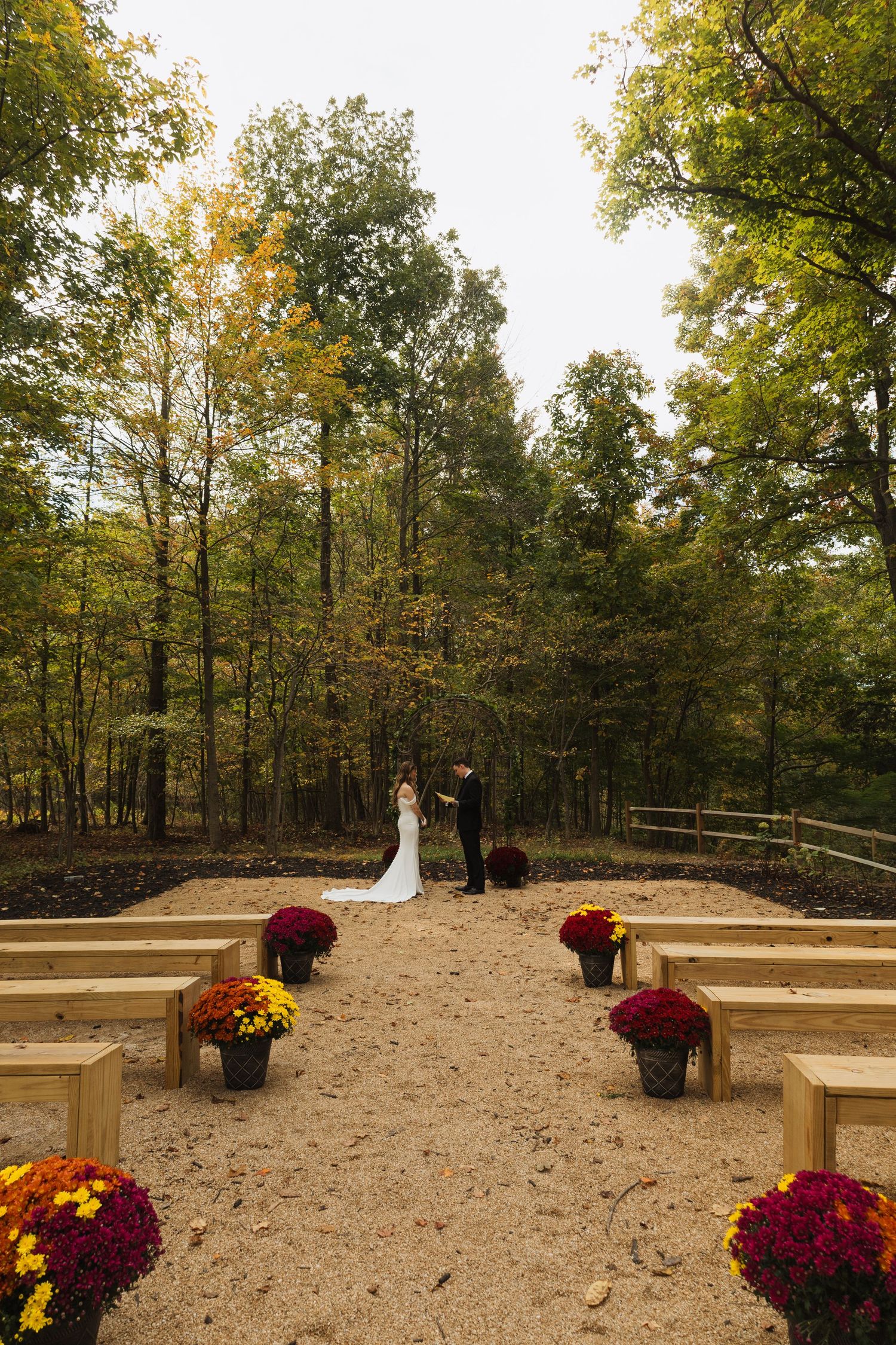 Outdoor wedding ceremony site with wooden benches and fall mums lining sandy aisle.