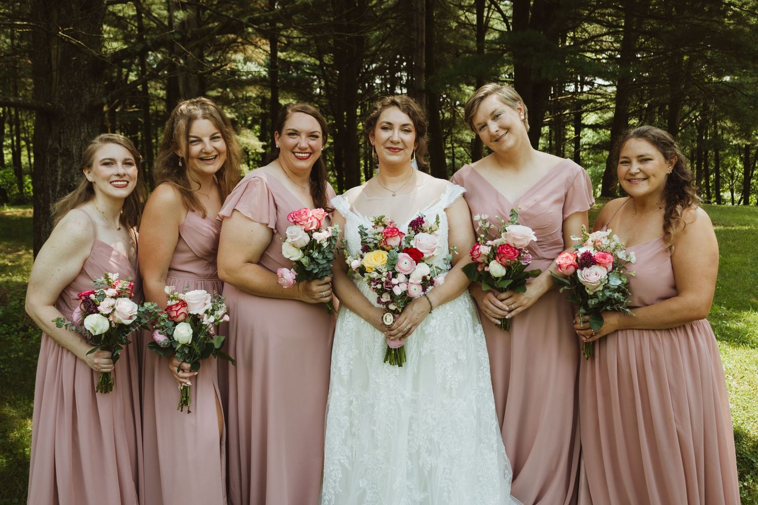Bridesmaids in dusty rose dresses pose with bouquets in an outdoor garden setting.