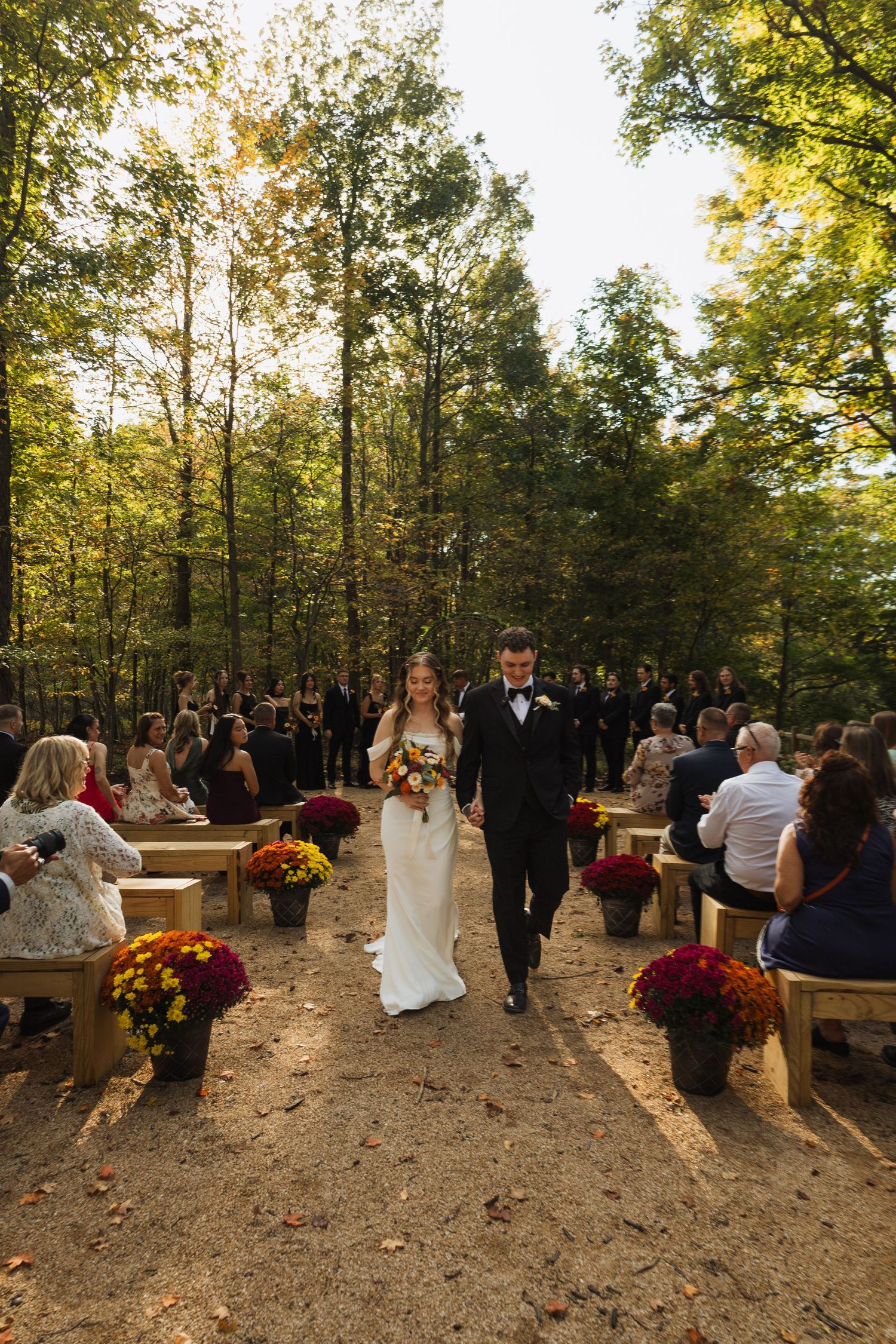 Outdoor autumn wedding ceremony in a wooded setting with wooden bench seating and fall decorations.