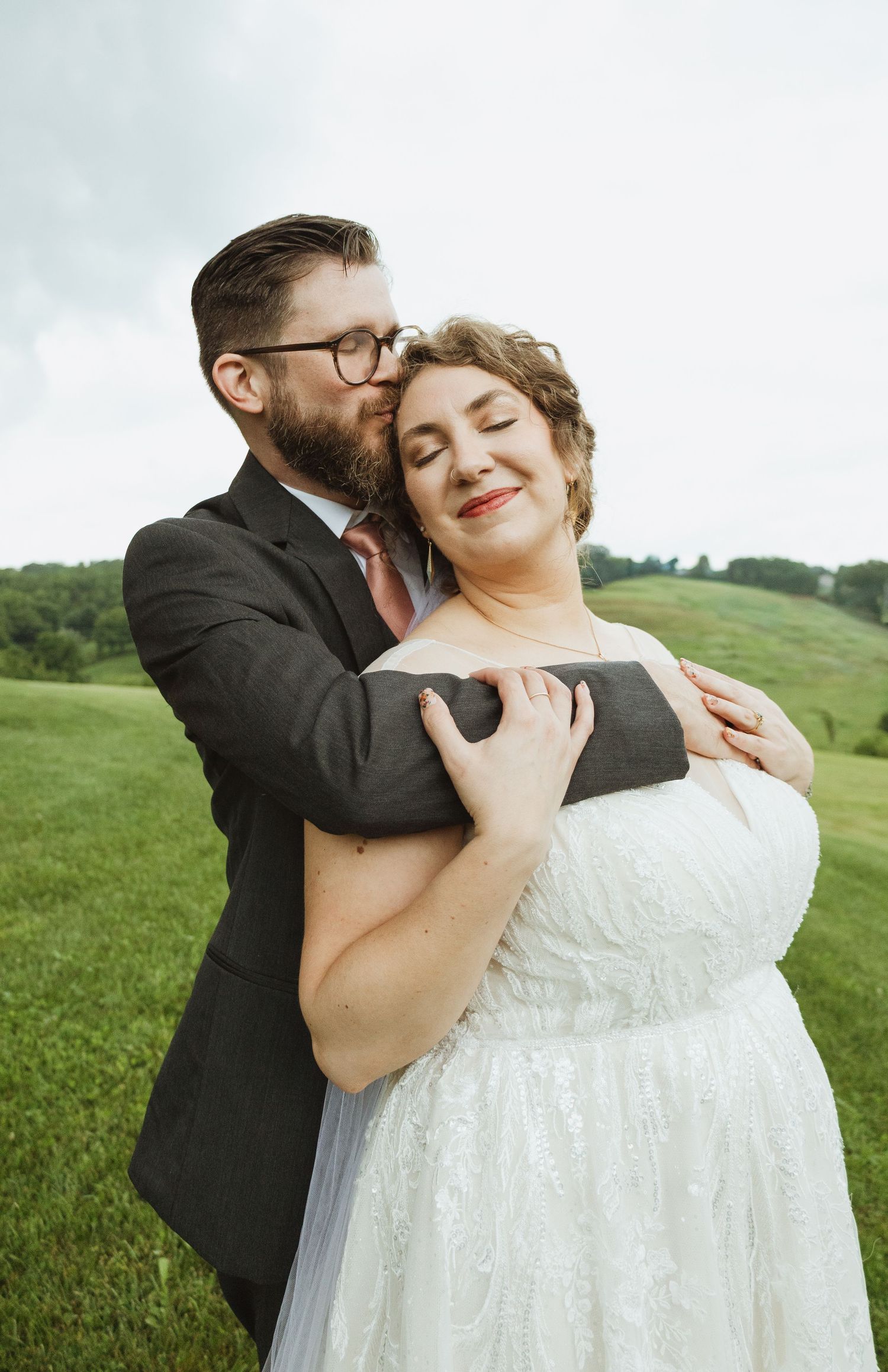 A romantic wedding portrait shows a couple in an intimate embrace against a lush green landscape.