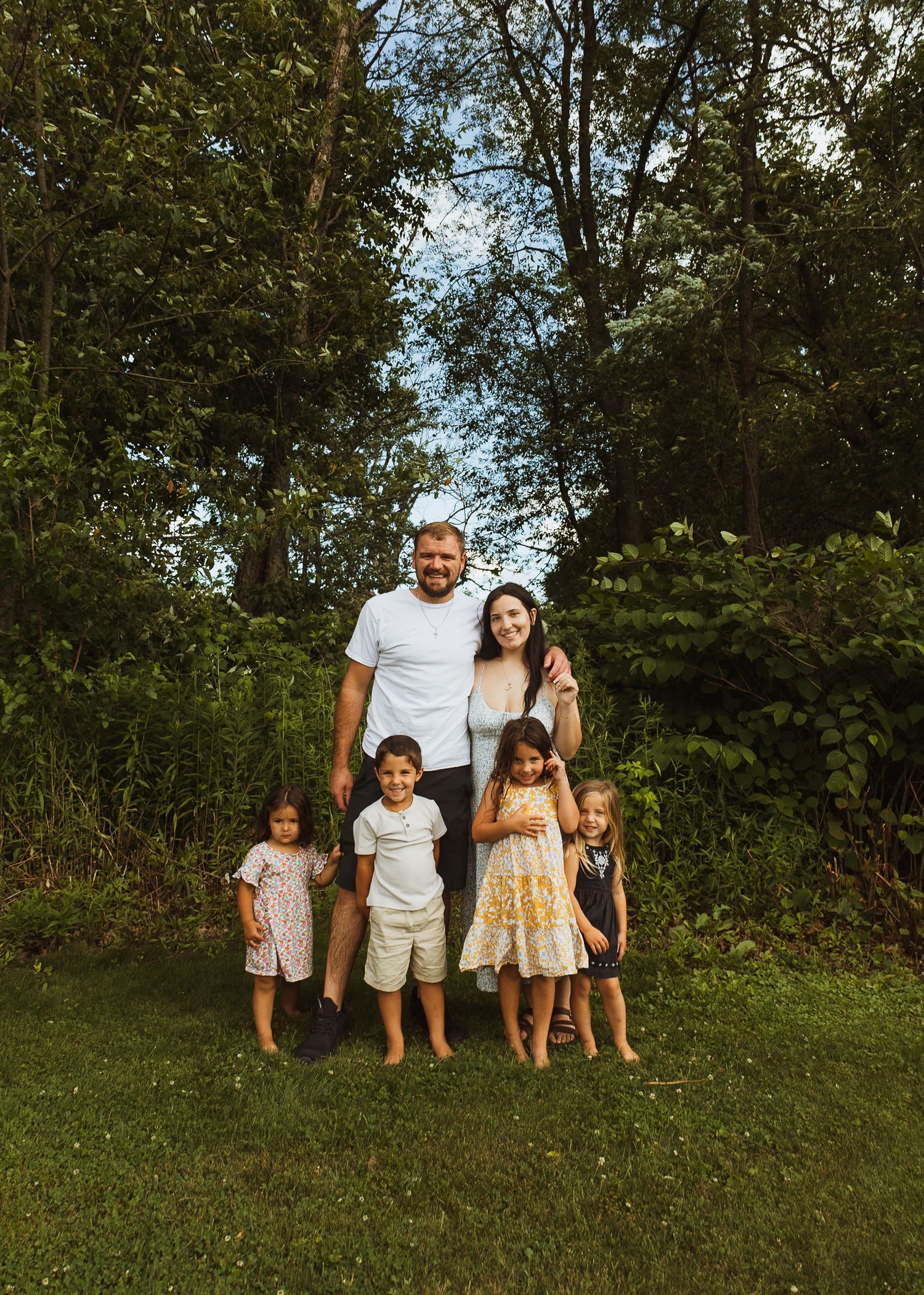Family of six posing outdoors on green lawn surrounded by lush trees on a warm summer day.