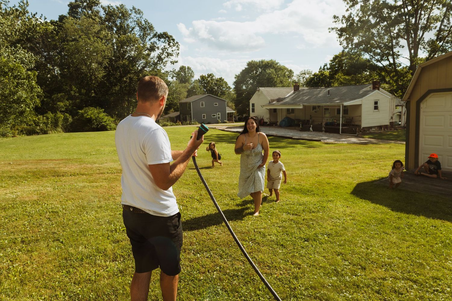 Man filming woman and child playing in sunny backyard with green lawn and suburban homes in background.