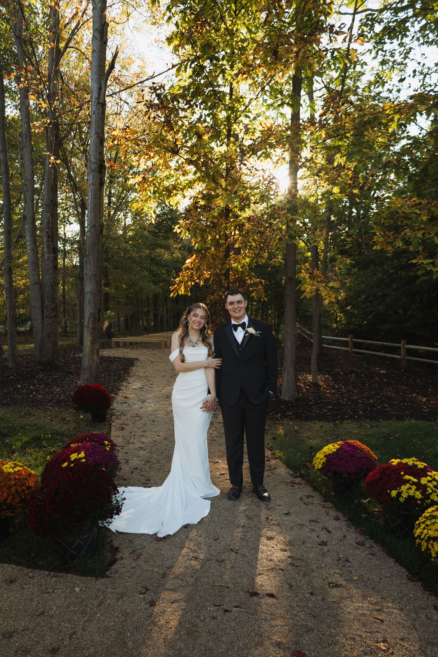 A wedding couple stands on a sunlit autumn path lined with red and yellow mums and tall trees.