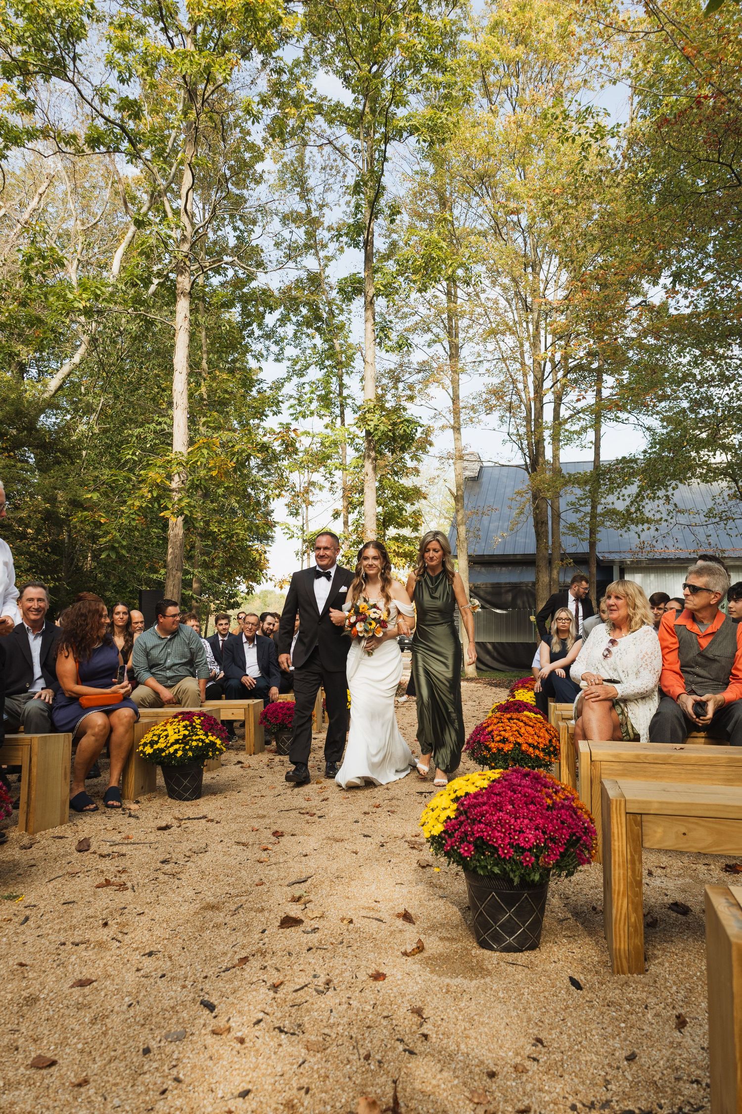 Wedding guests seated on wooden benches at an outdoor ceremony venue surrounded by fall foliage.