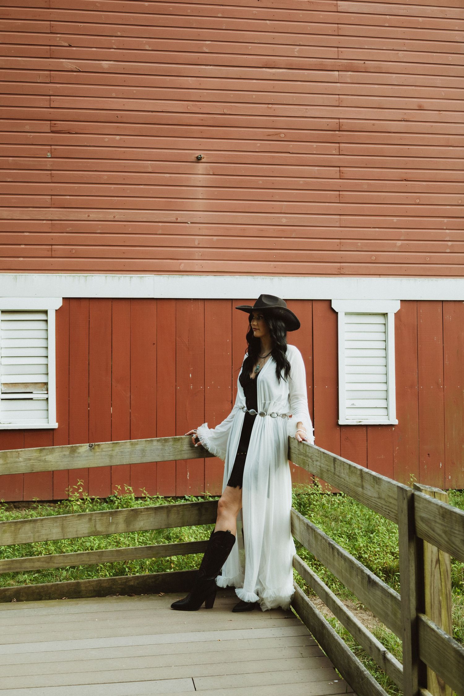 A person in a flowing white dress stands on a rustic wooden walkway next to a red barn wall.