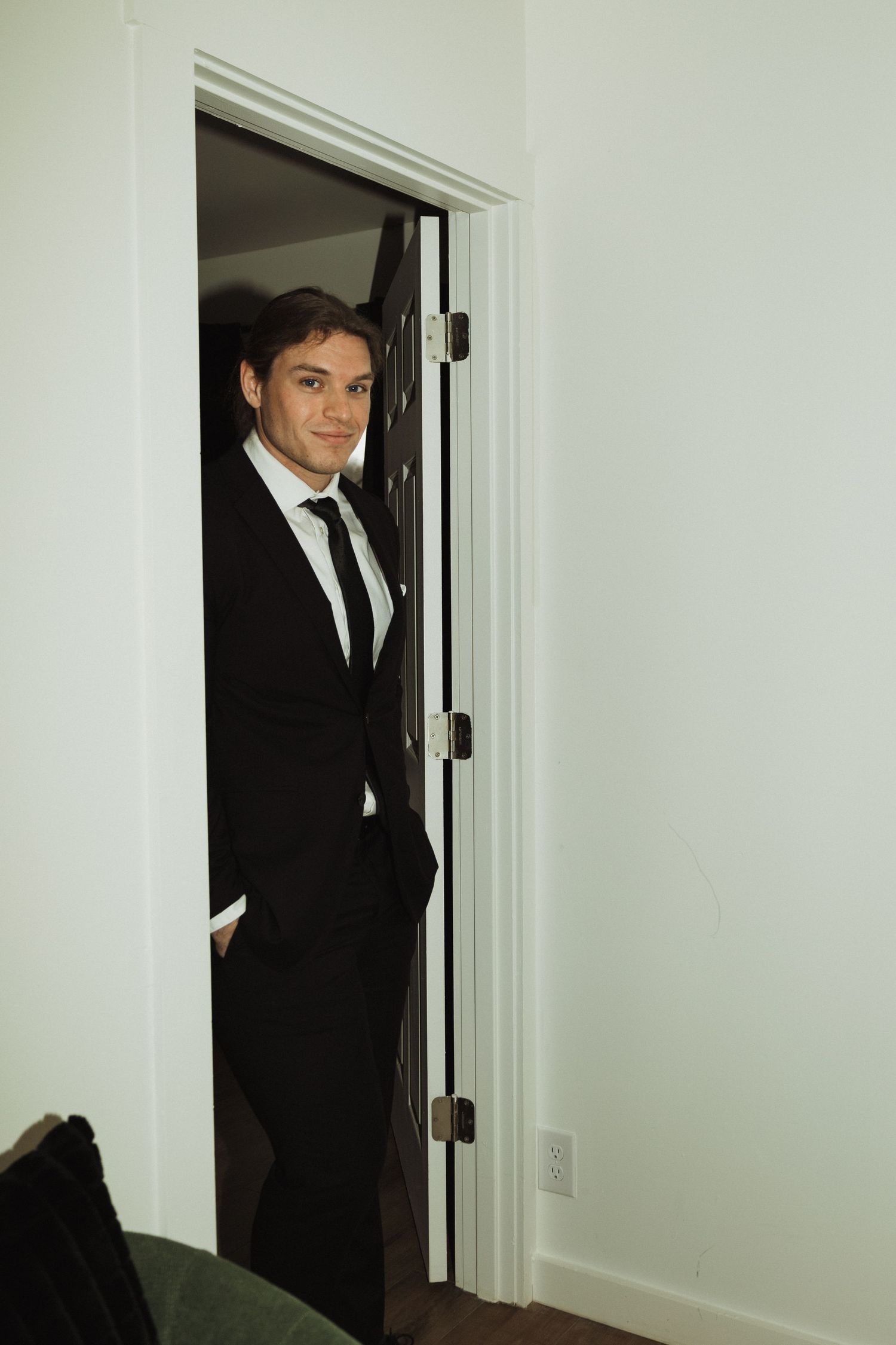 Person poses confidently in doorframe wearing classic black suit and tie.