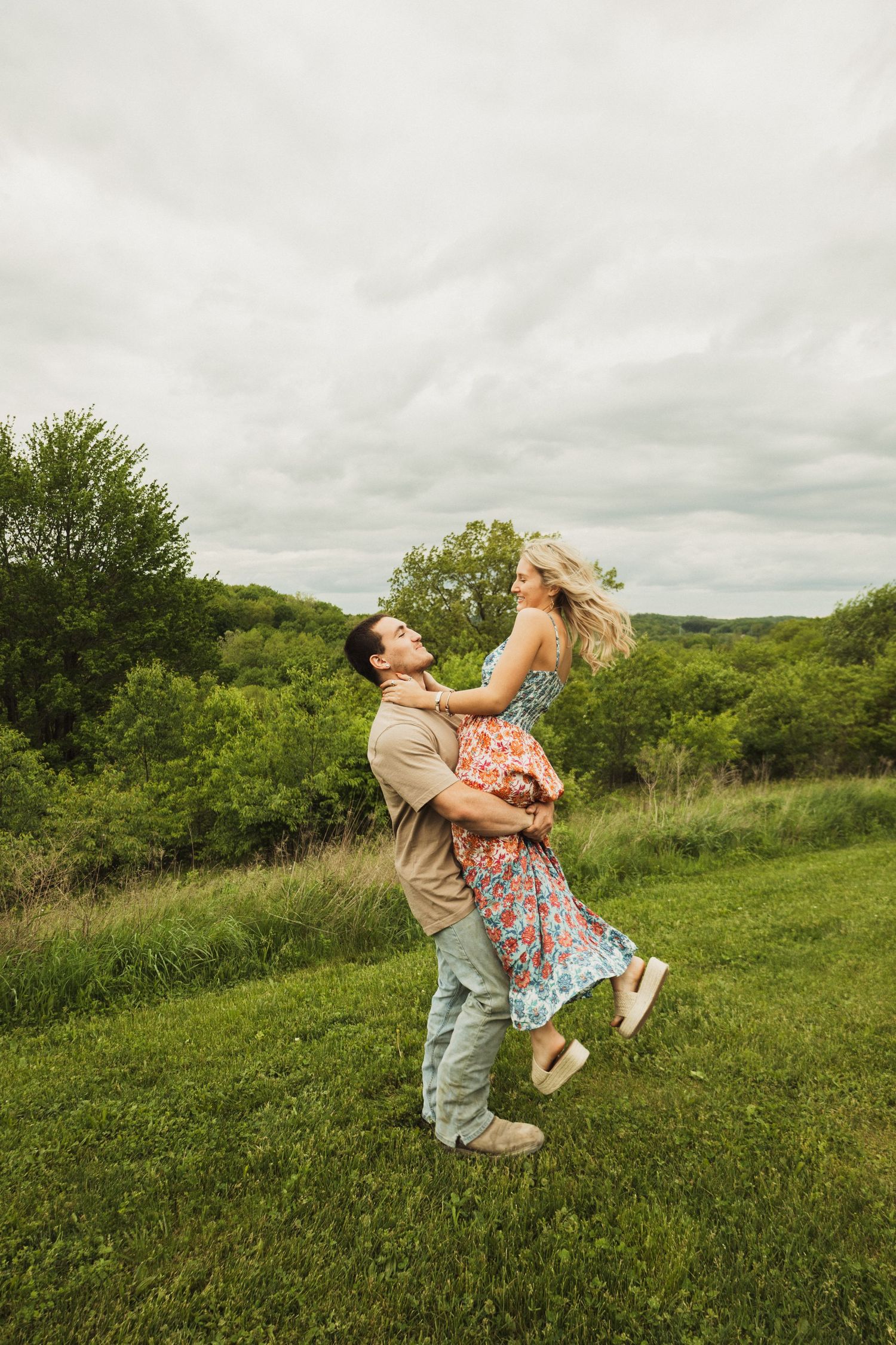 A couple share a joyful moment dancing and lifting each other in a green hillside meadow on an overcast day.