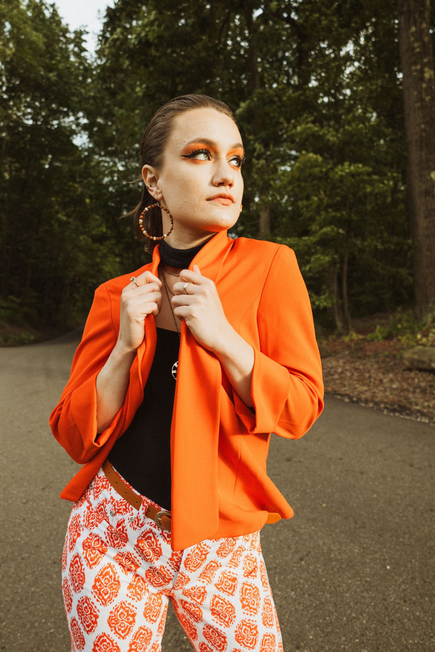 Fashion model in vibrant orange blazer and patterned pants poses on outdoor path with trees in background.