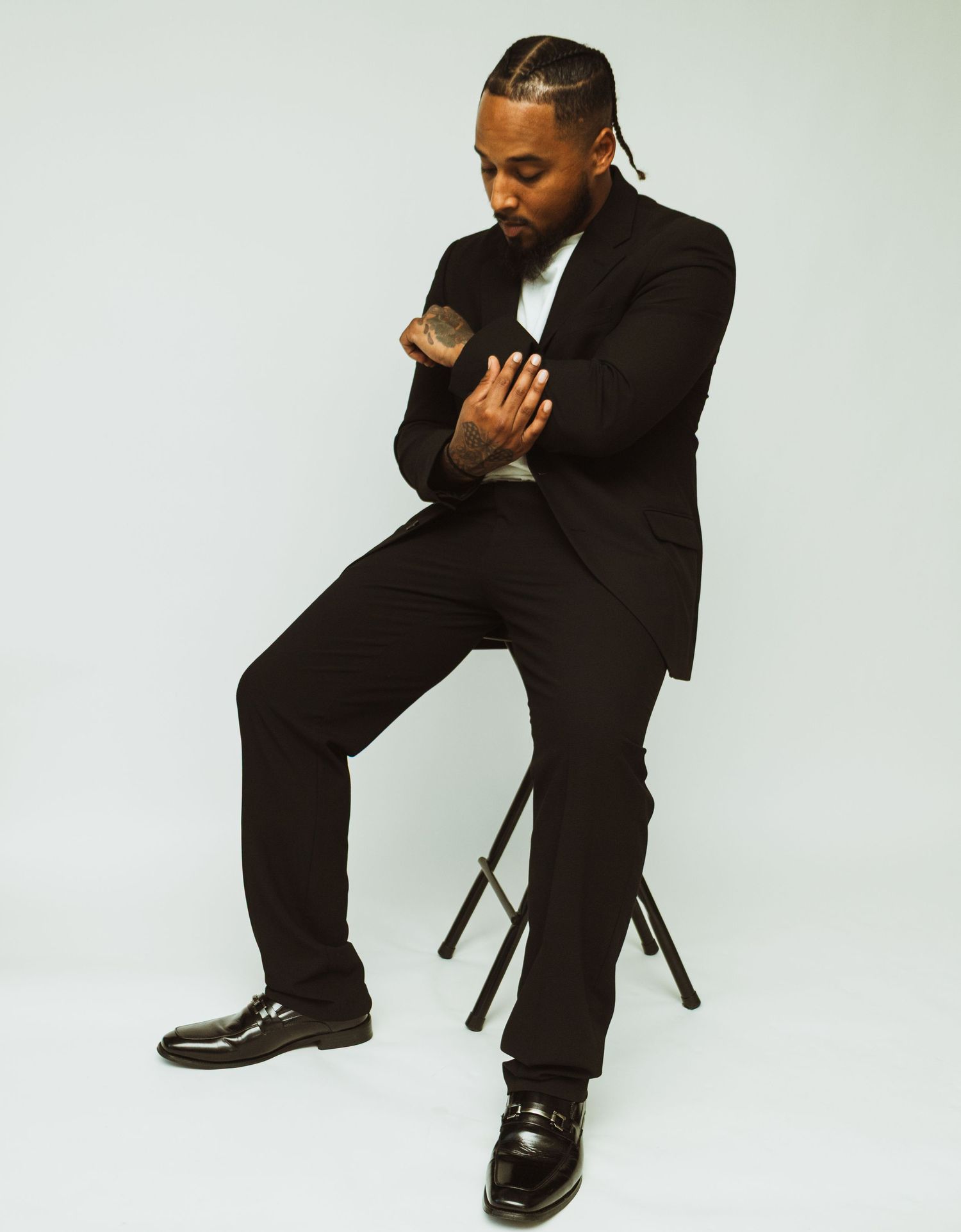 Person in black suit sitting on stool in artistic studio portrait against light background.