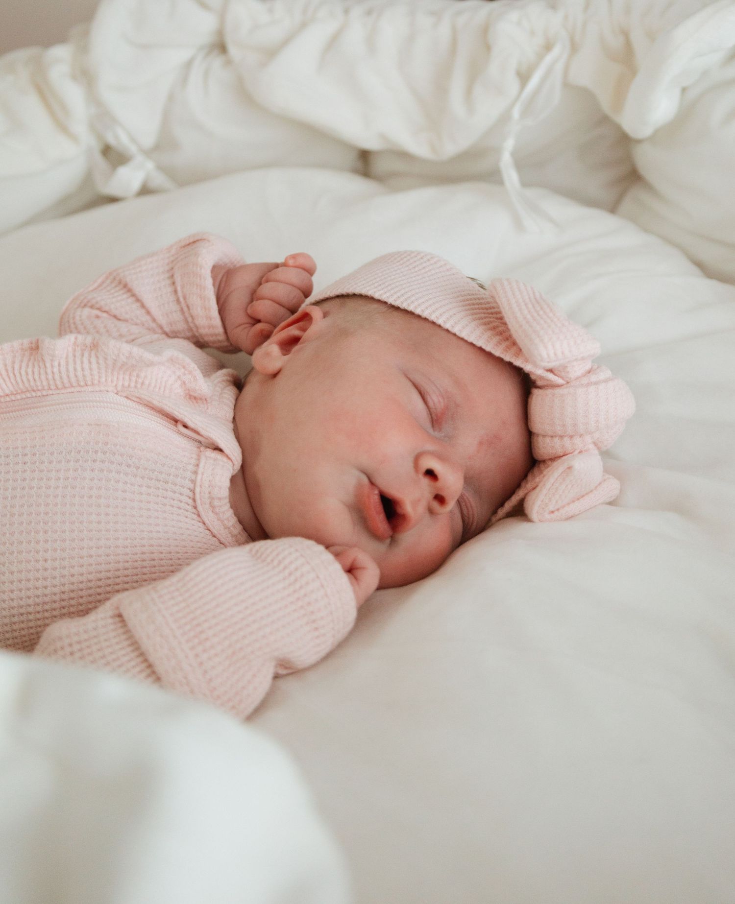 Sleeping baby wearing soft pink knit sweater rests peacefully on white bedding.