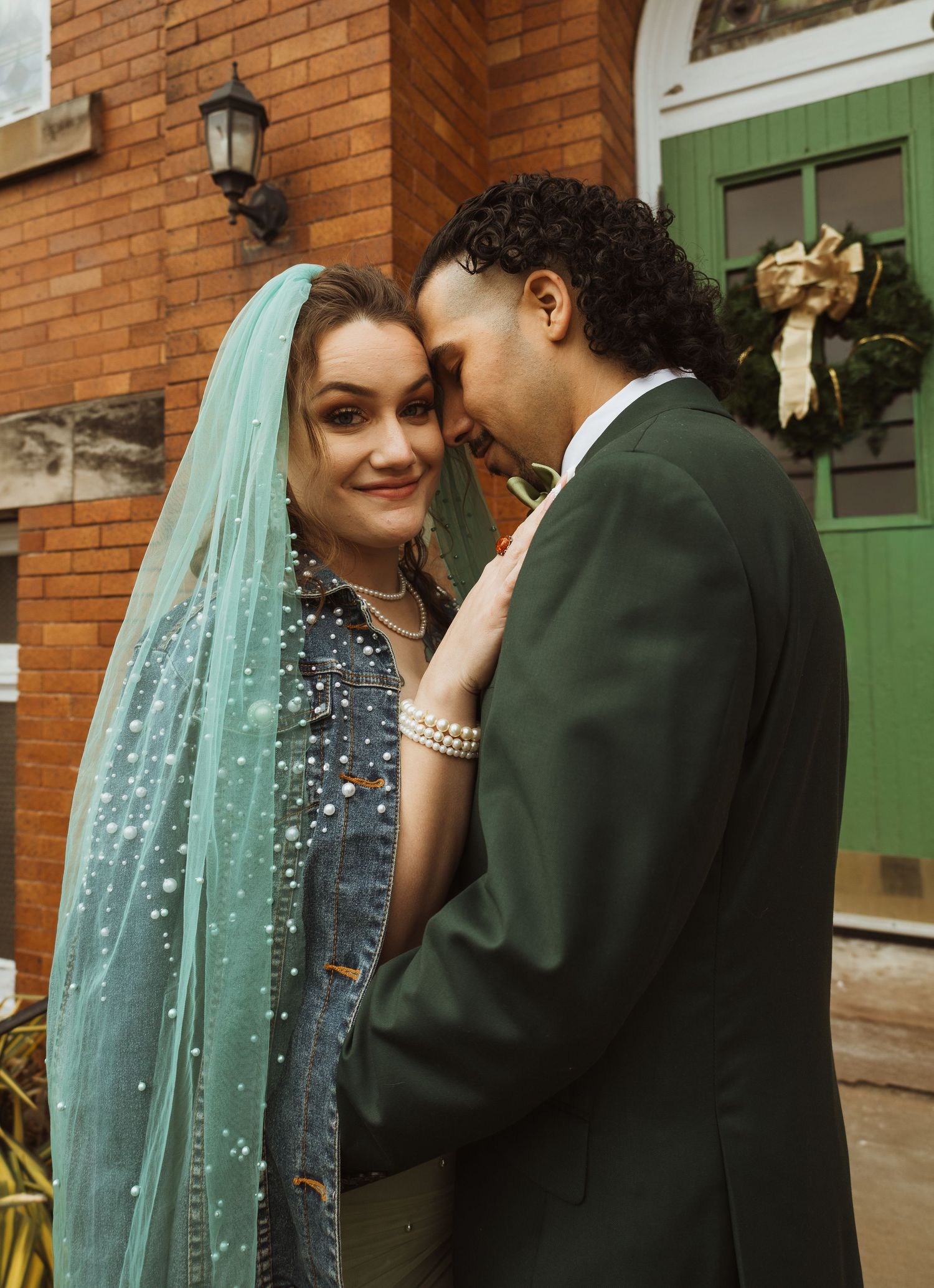 Smiling bride in teal veil and denim jacket with groom in green suit outside brick building.
