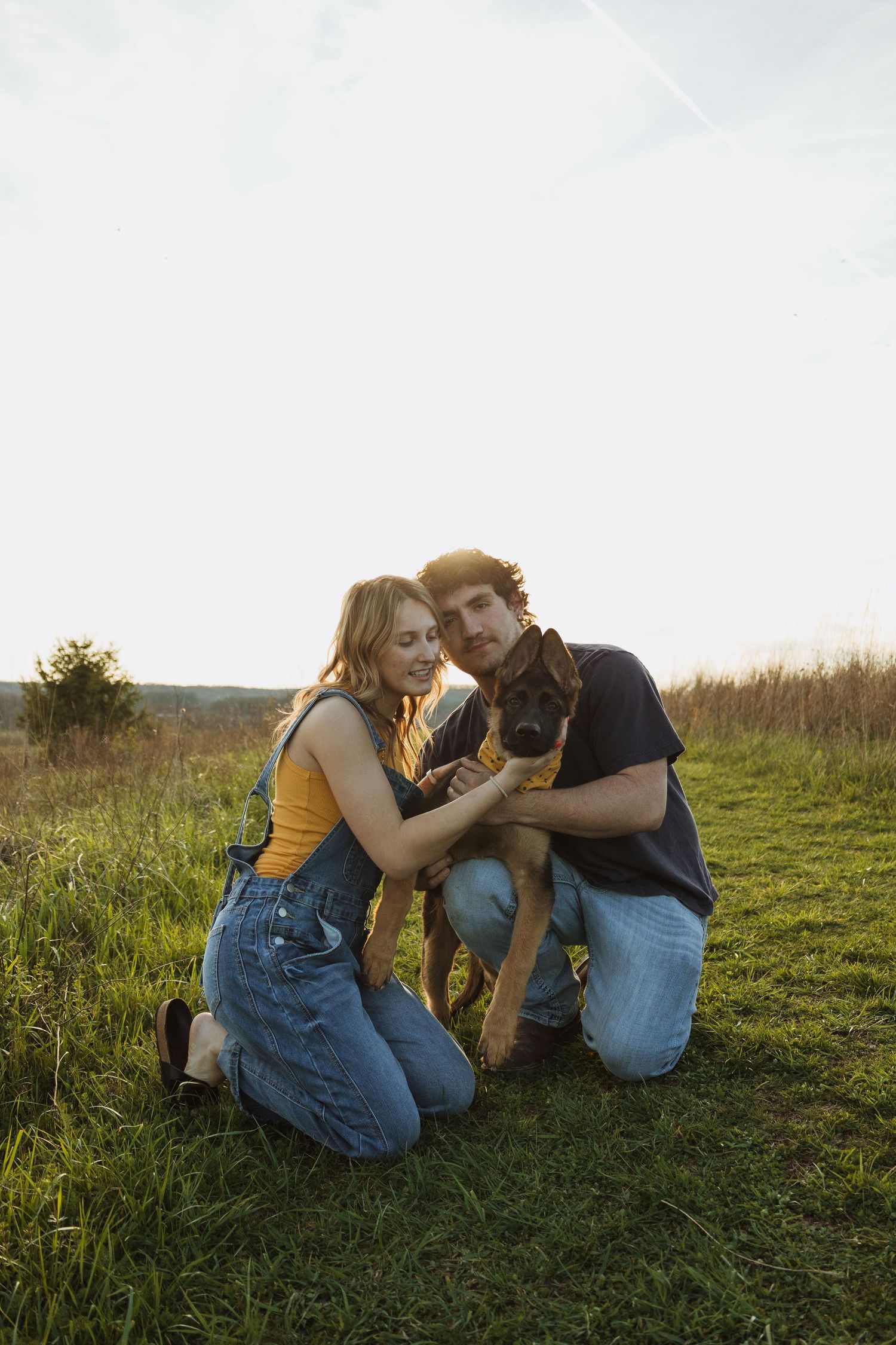 A couple kneels in a grassy field at sunset, lovingly petting their brown dog together.