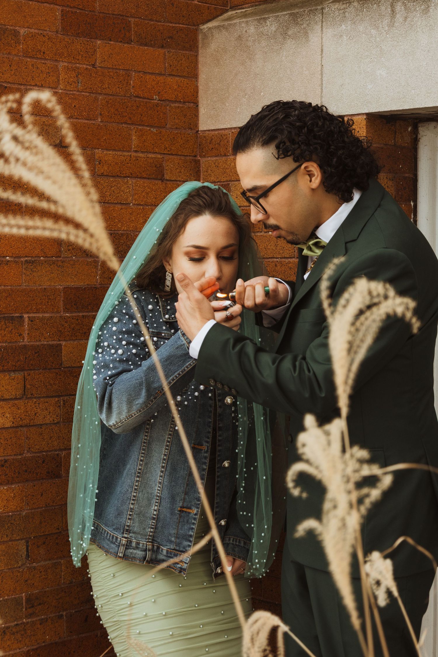 Groom in dark green suit lights bride's cigarette outdoors near brick wall with pampas grass in foreground.