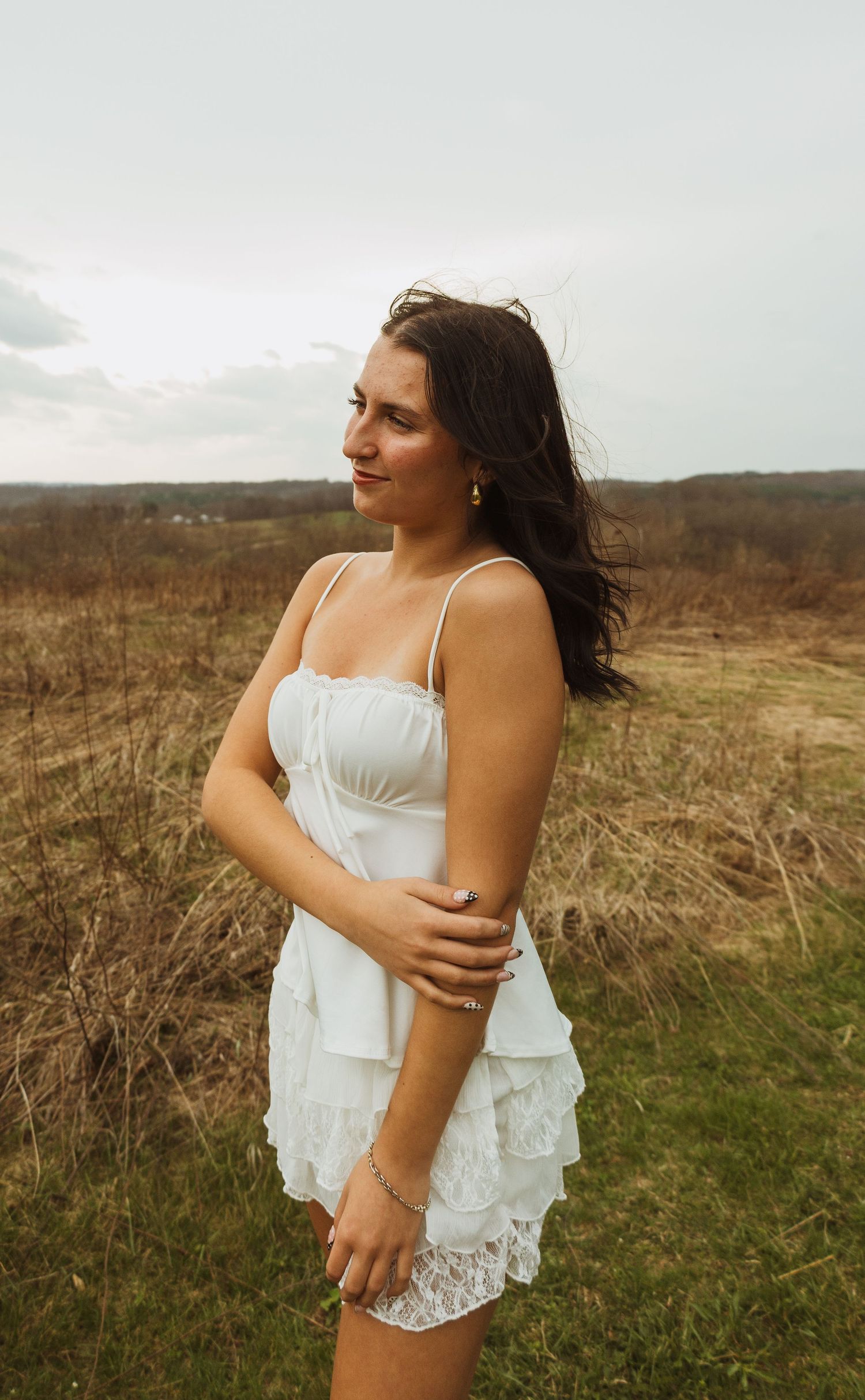 Young woman in white ruffled dress standing in a field, hair blowing in the wind on an overcast day.