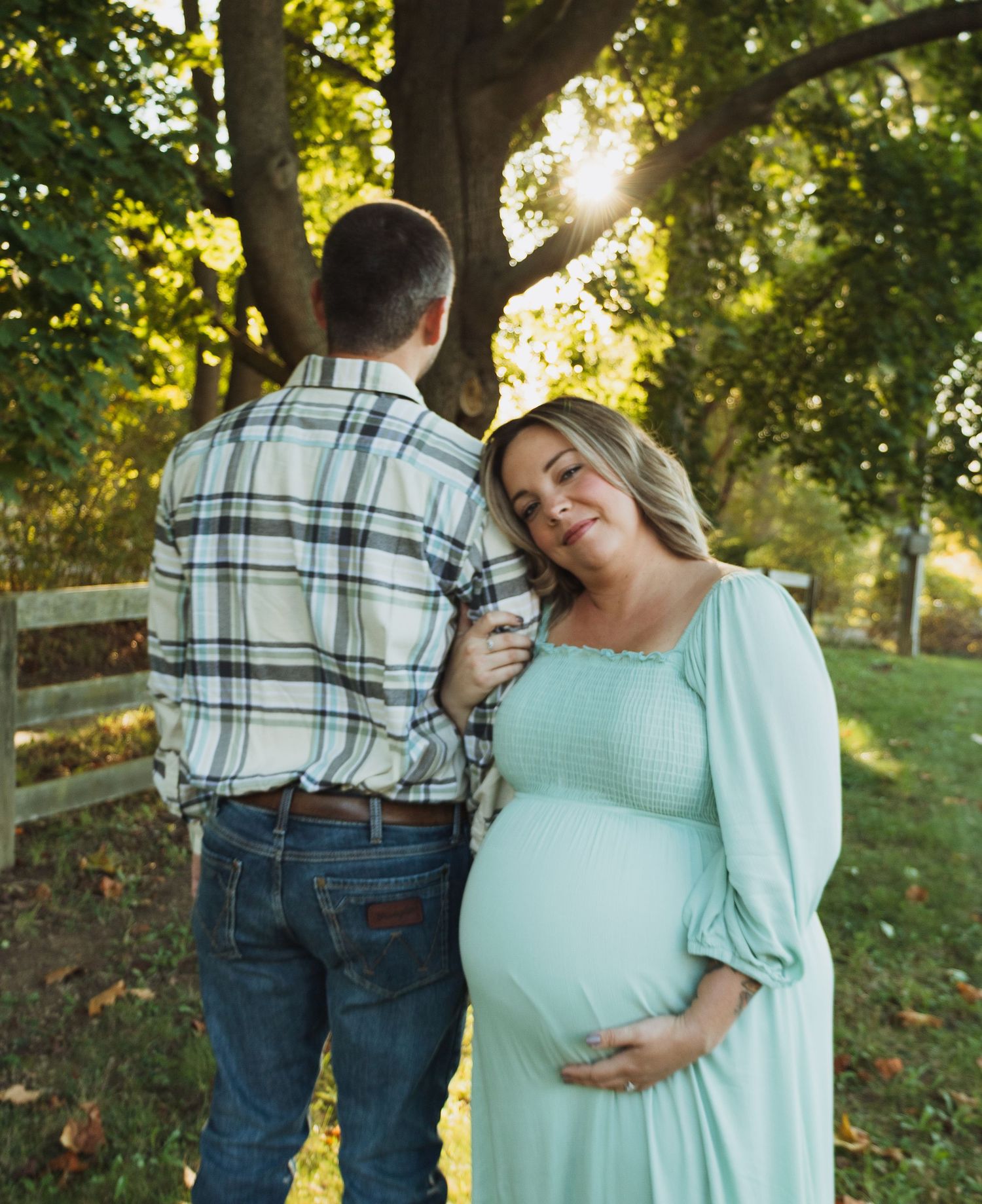A couple embraces in a sun-dappled wooded area during a maternity photo session, wearing mint green and plaid.