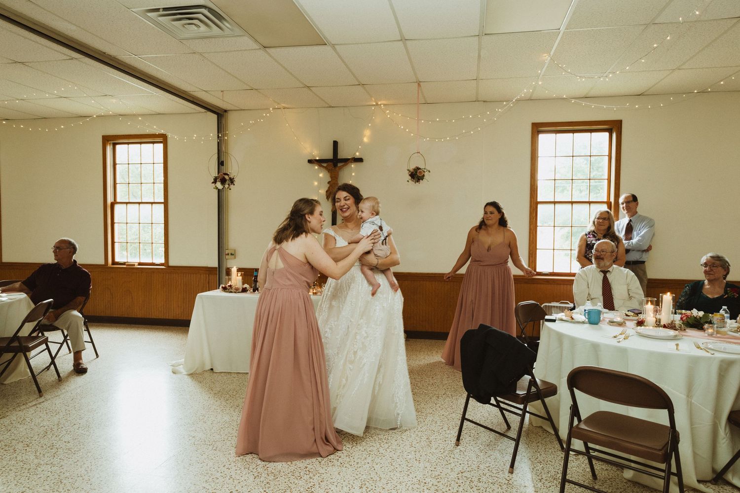 Wedding reception in a church hall with guests seated at round tables and string lights overhead.