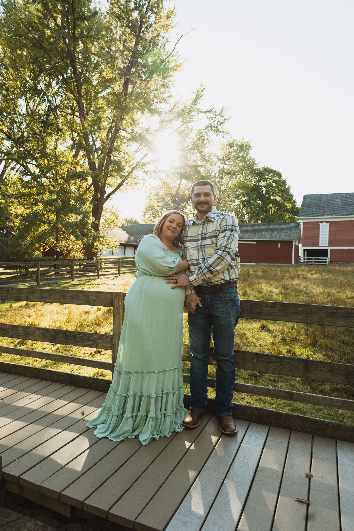 Couple shares tender moment on wooden deck with sunlit trees and red barn in background during golden hour.