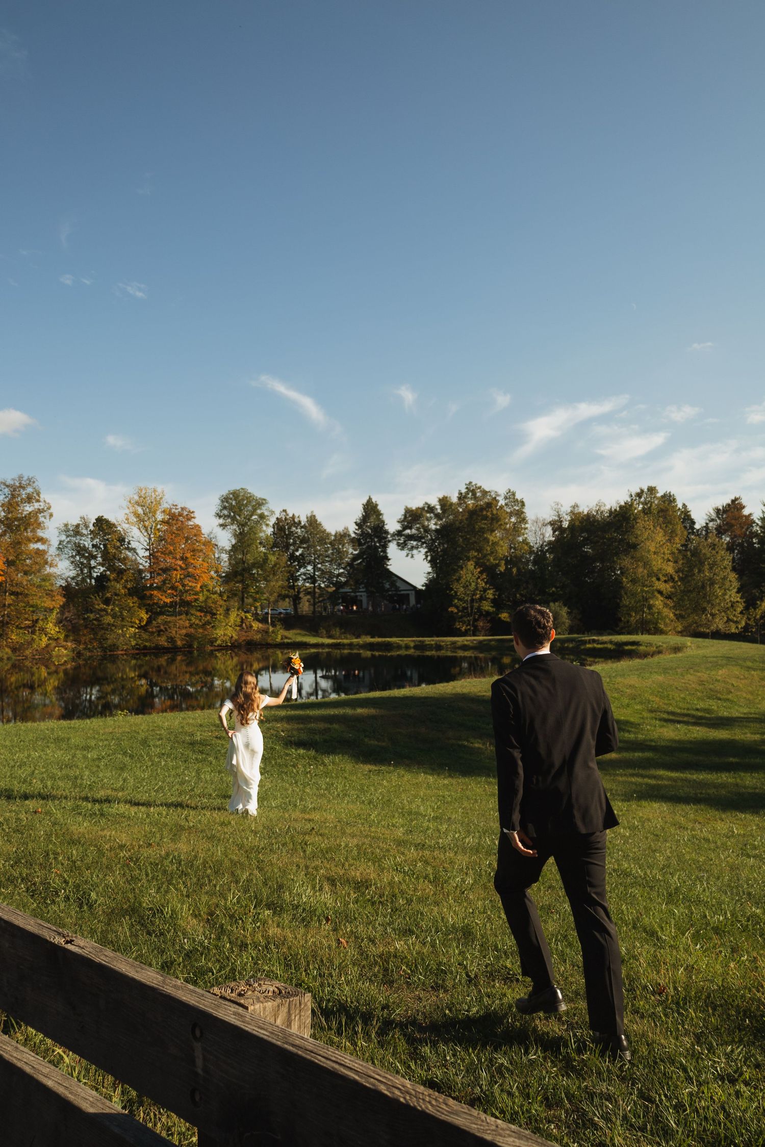 Couple in wedding attire standing apart on grassy hill during autumn sunset.
