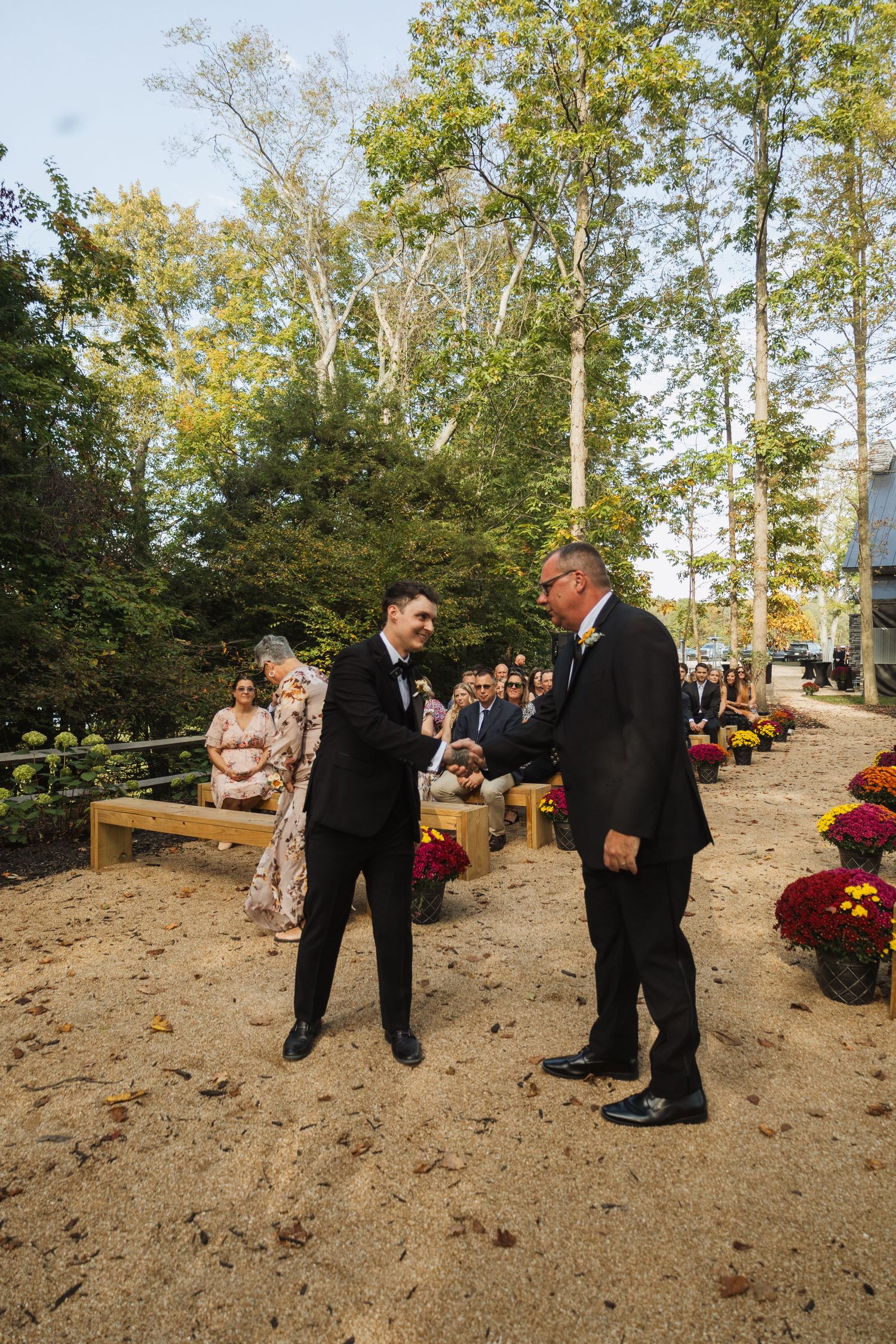 A formal exchange during an outdoor autumn wedding ceremony with guests seated on wooden benches.