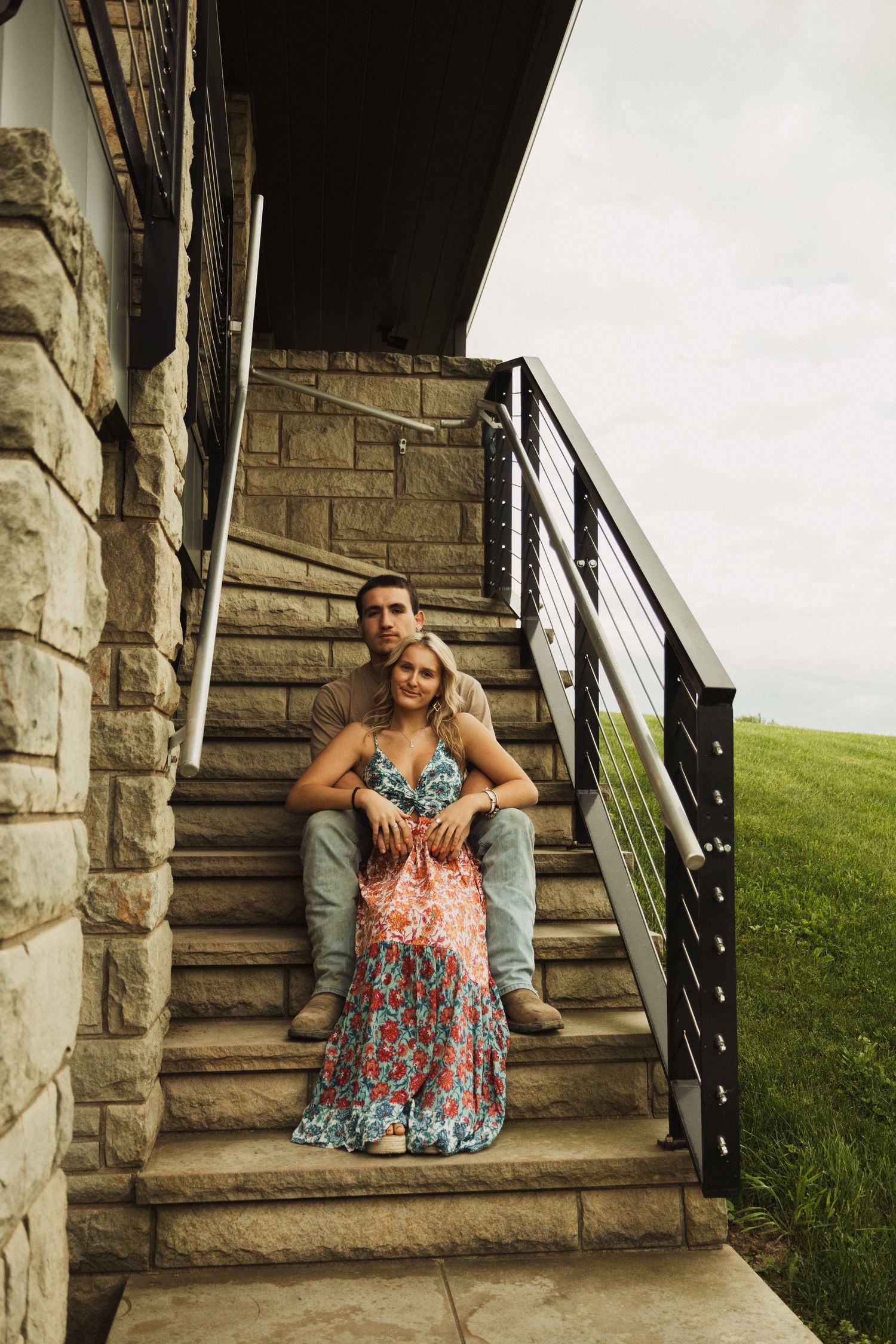 A couple sits together on stone stairs with metal railings beside a grassy hill during golden hour.