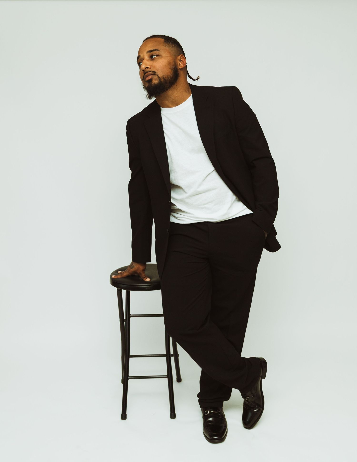 A person in a black suit and white t-shirt poses casually while leaning against a tall bar stool on a light background.