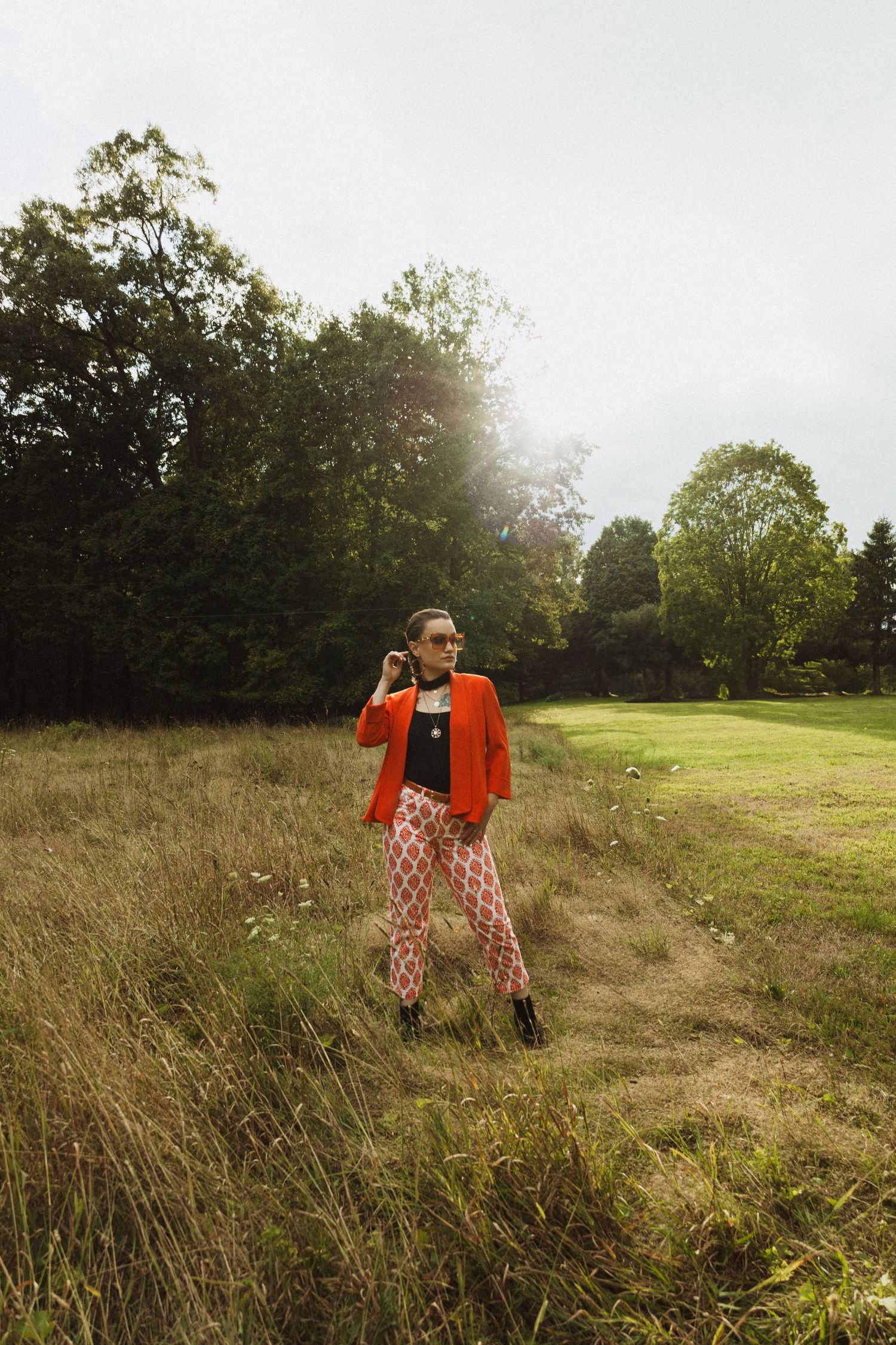 Candid fashion shot featuring bold orange blazer and printed pants against rural landscape.