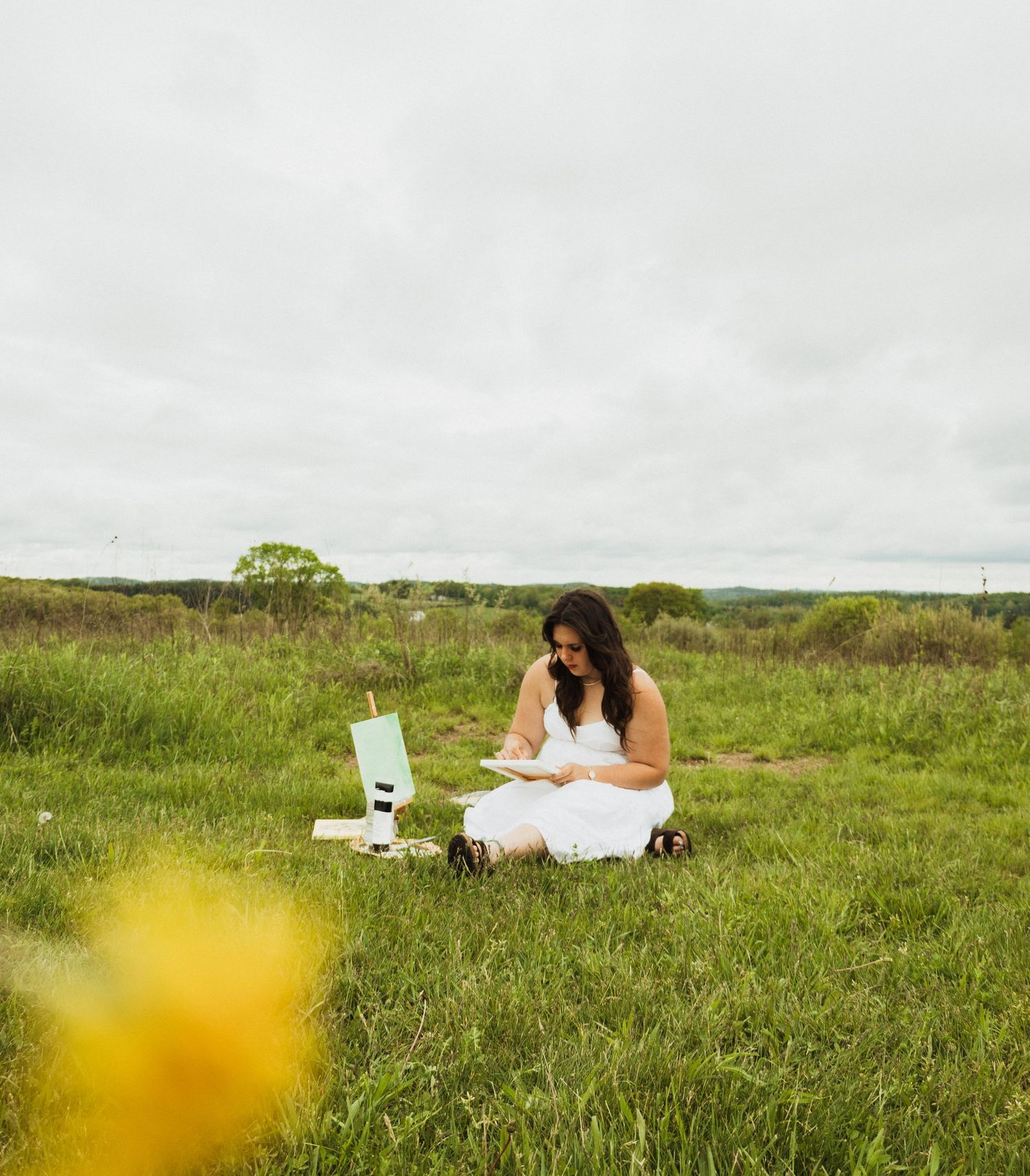 A person in a white dress sits in a grassy meadow with a laptop and drink, enjoying nature on an overcast day.