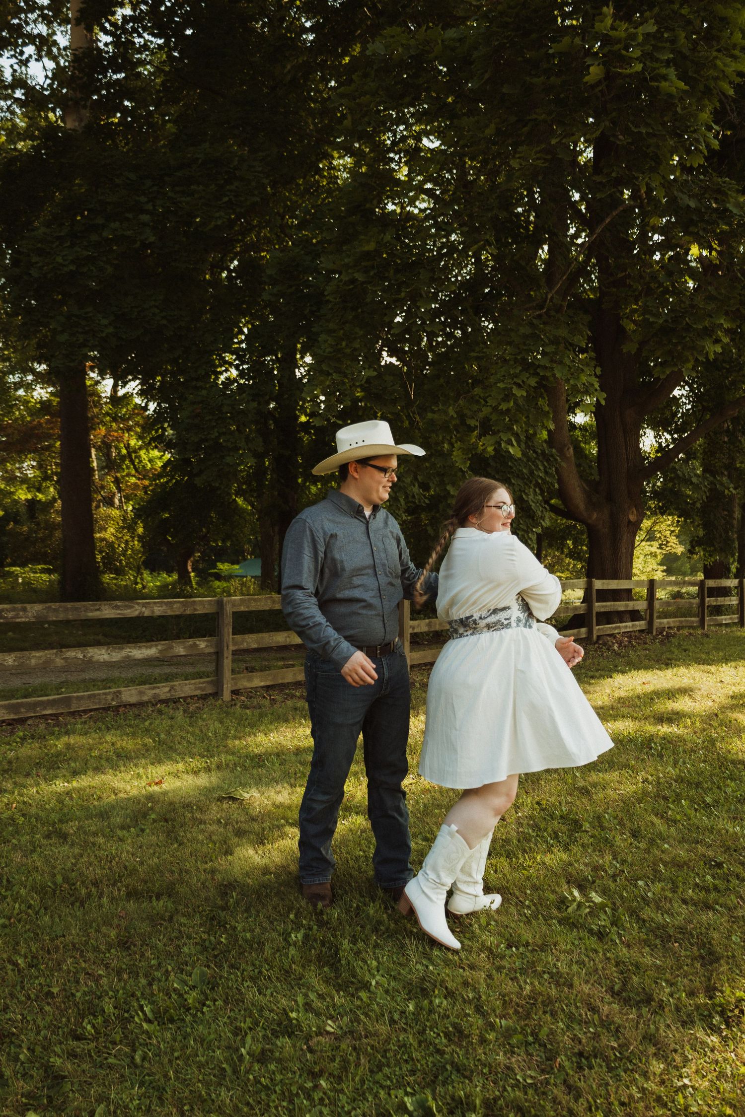 A couple dances together on a grassy lawn near a rustic wooden fence during golden hour.