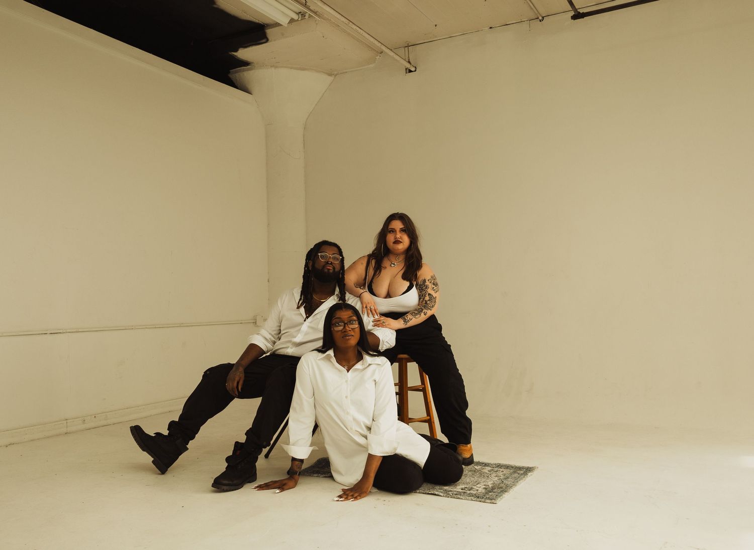Three people in contrasting black and white outfits pose casually together on a gray rug in a white studio space.