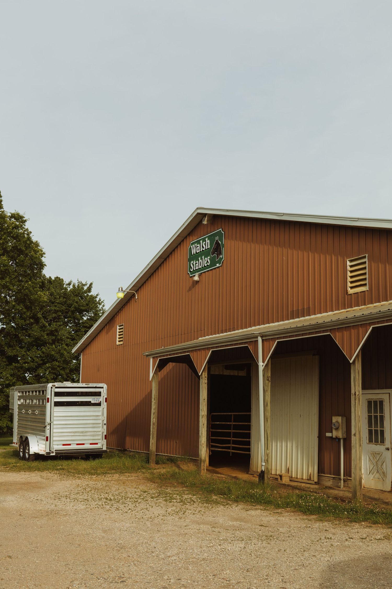 Red barn with Welsh Stables sign, white livestock trailer parked outside on a rural gravel property.