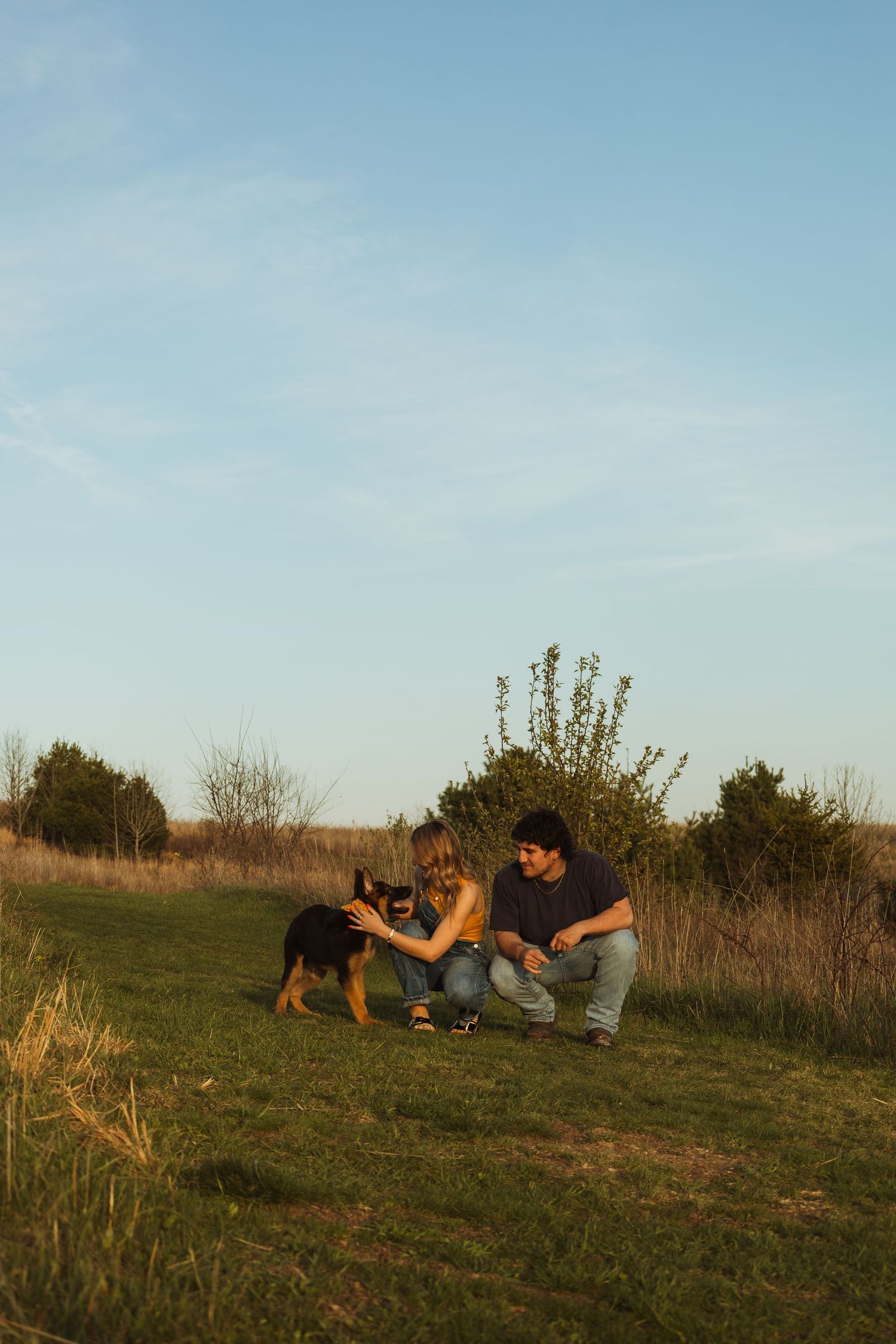 Couple crouching on green grass petting a German Shepherd puppy outdoors under a clear blue sky at sunset.