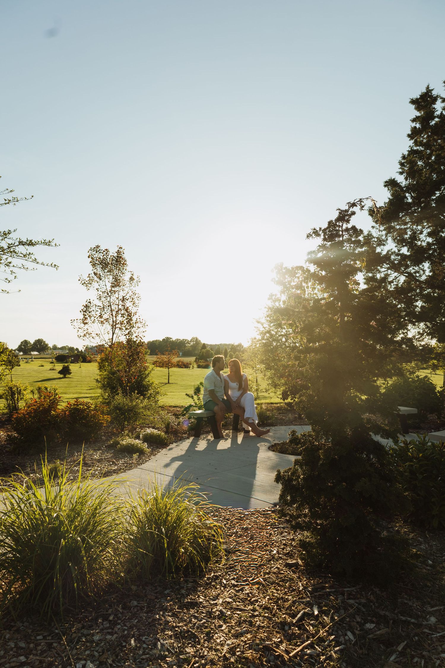 Couple standing on a garden path at golden hour, surrounded by lush greenery and trees with sunlight glowing behind them.