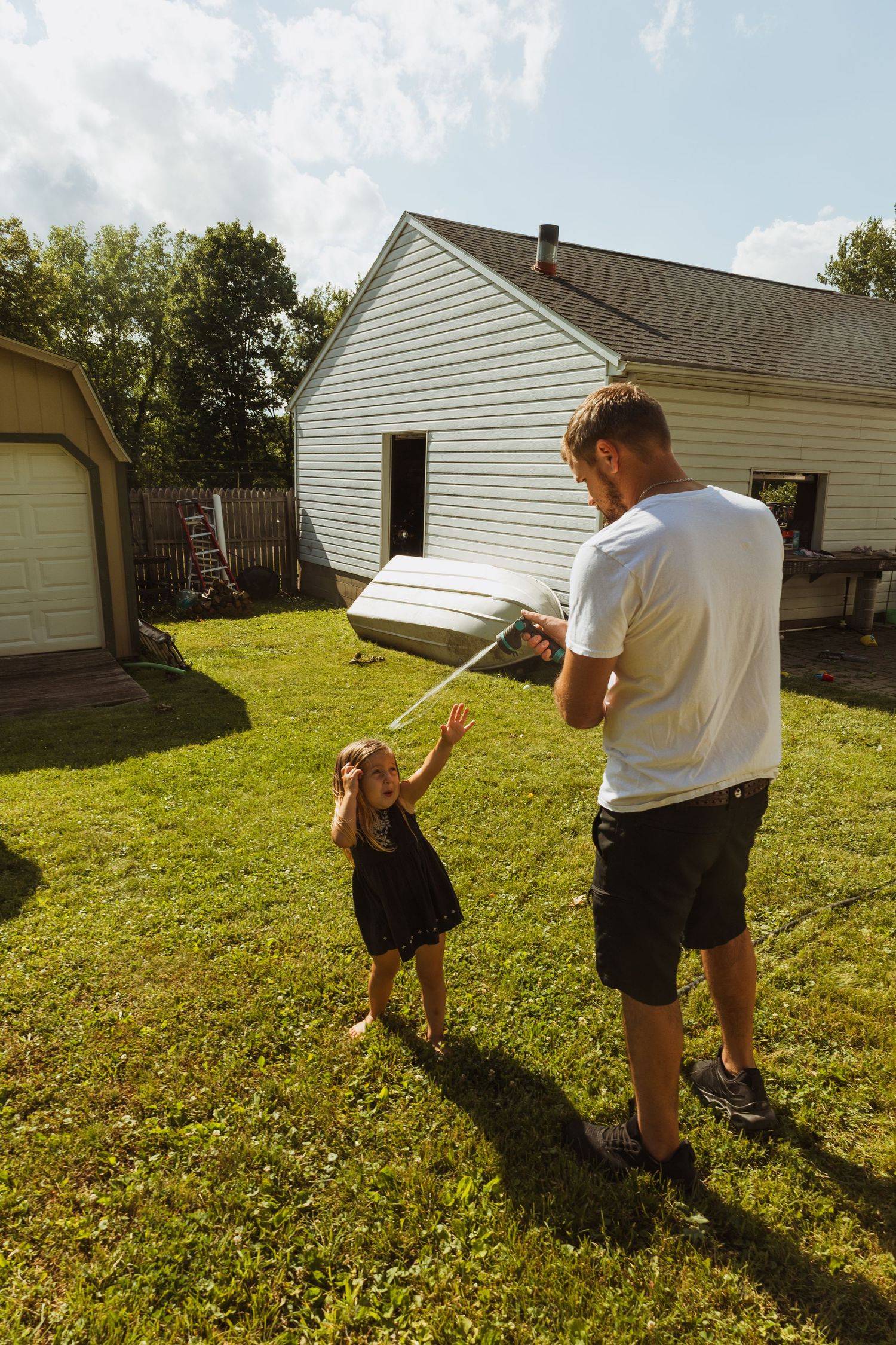 Father and young child playing in backyard on sunny day near white house with green lawn.