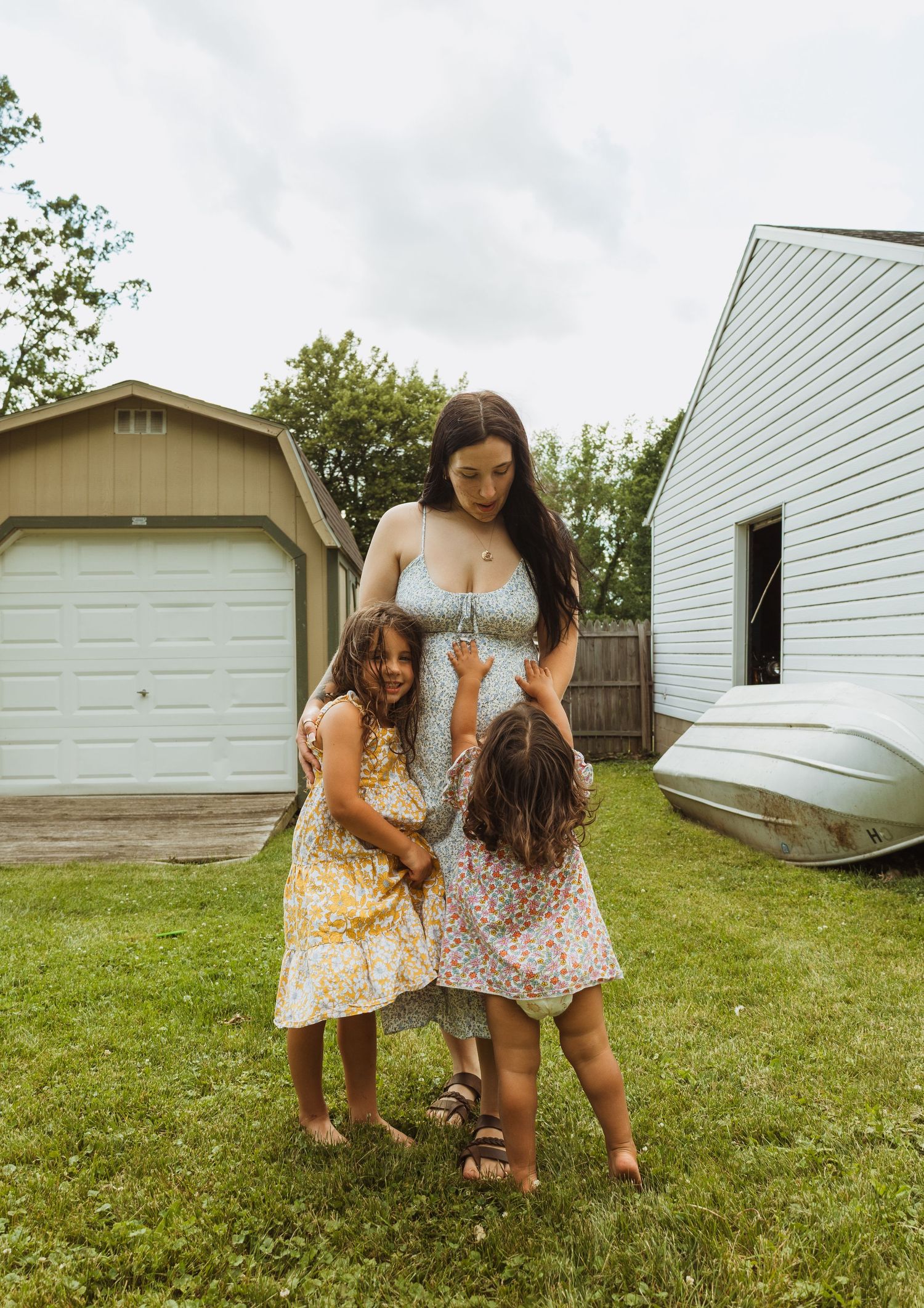 A pregnant woman in a light blue dress stands in a backyard with two young girls in floral dresses on a cloudy day.