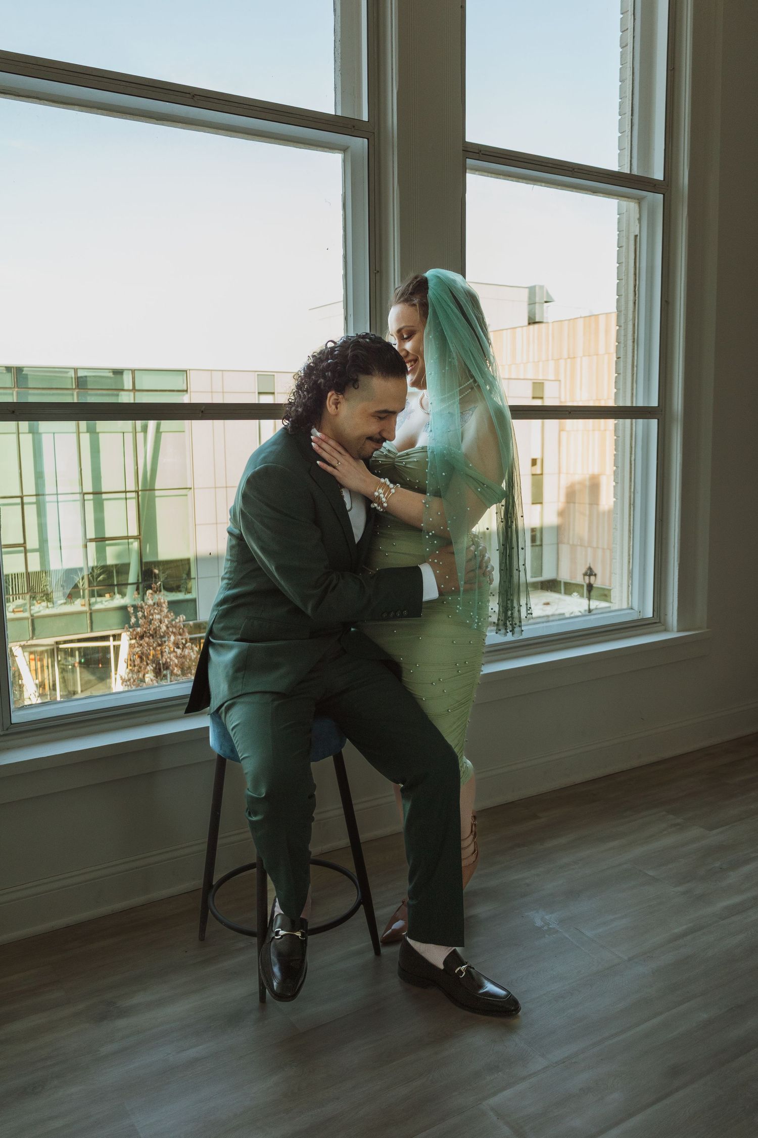 Bride in green veil embraces seated groom in dark green suit before large sunlit windows.