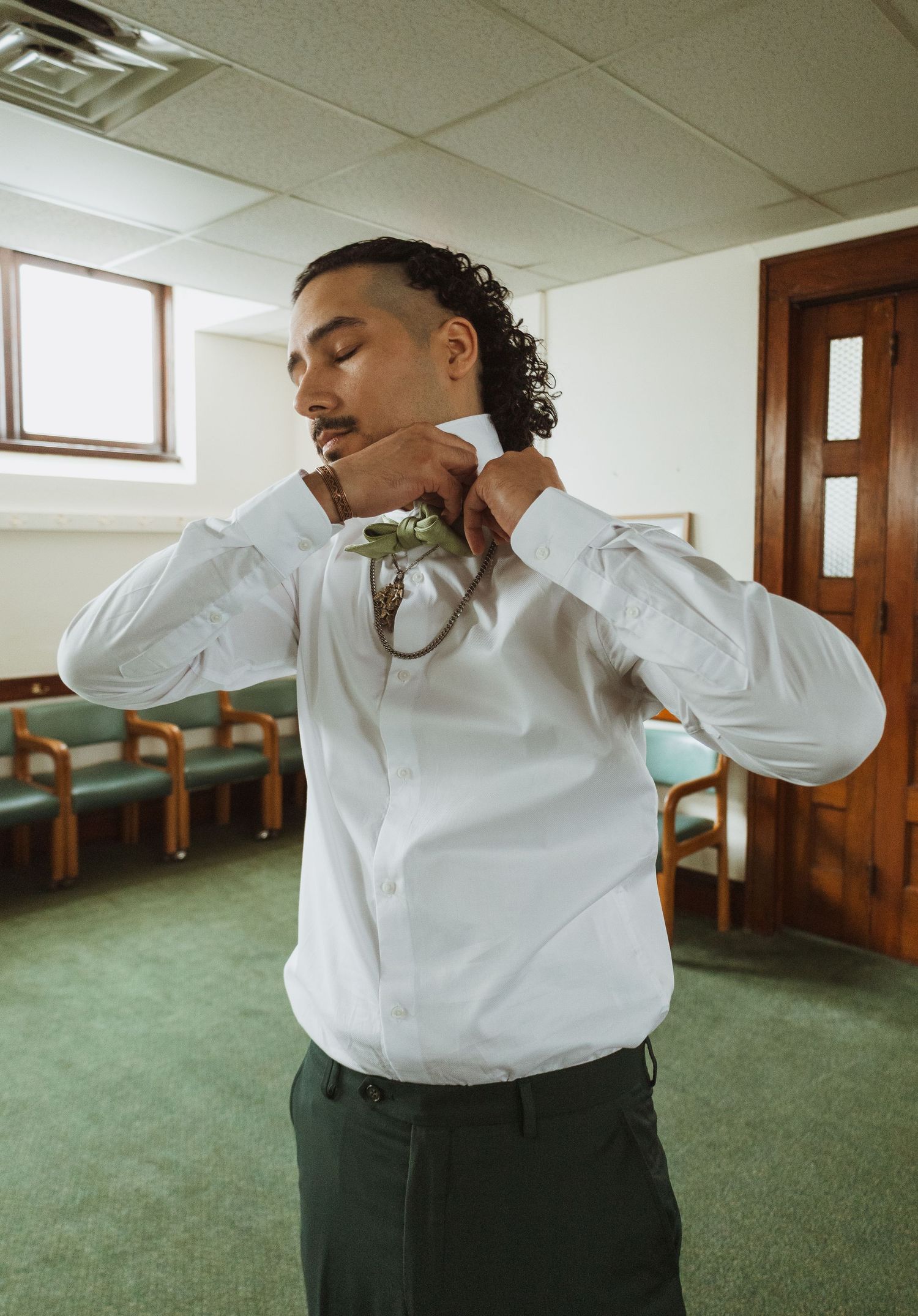 Groom in white dress shirt pinning green floral boutonniere while getting ready for wedding.