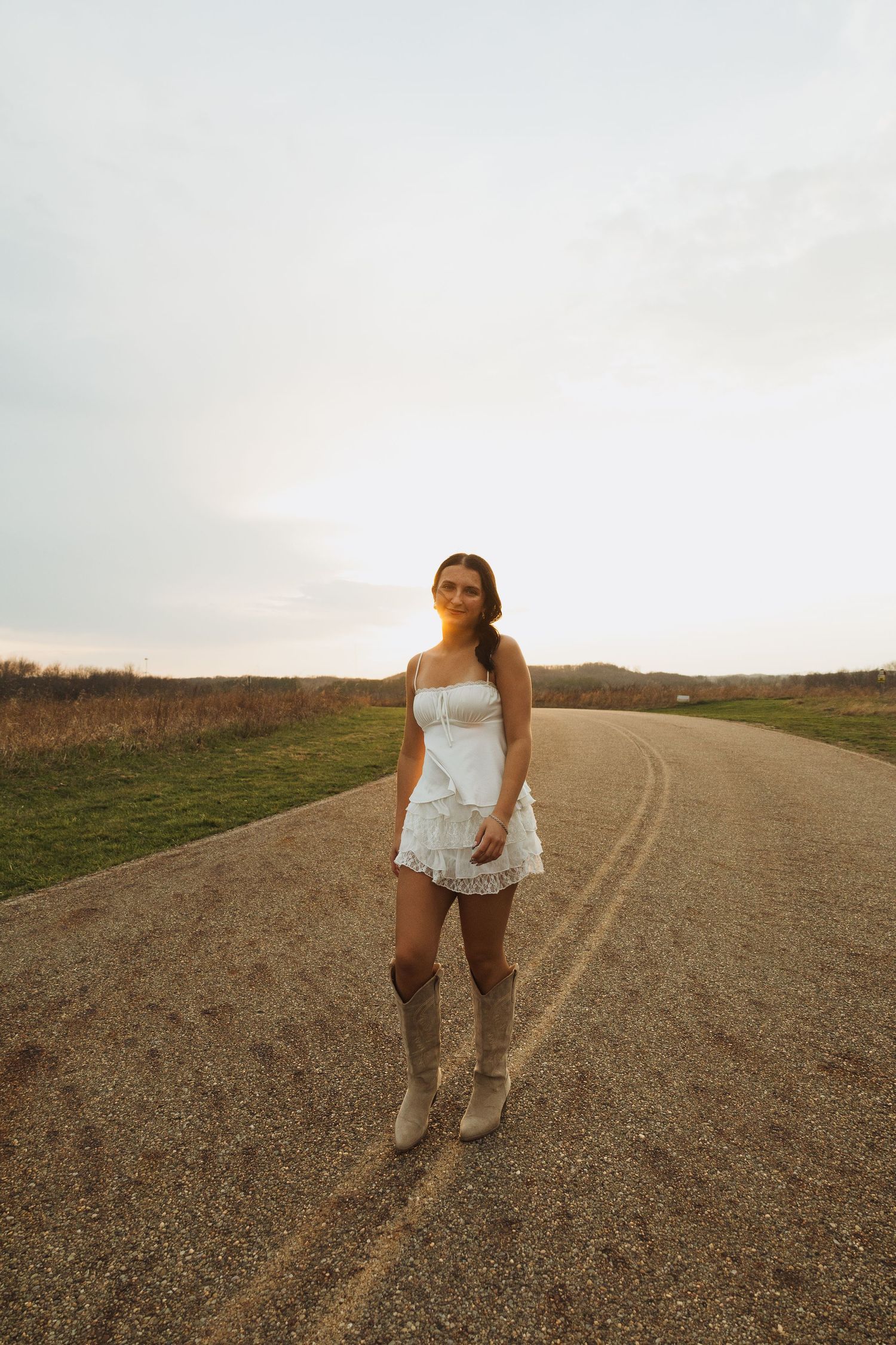 Young woman in white lace dress and cowboy boots standing on a rural dirt road at sunset.