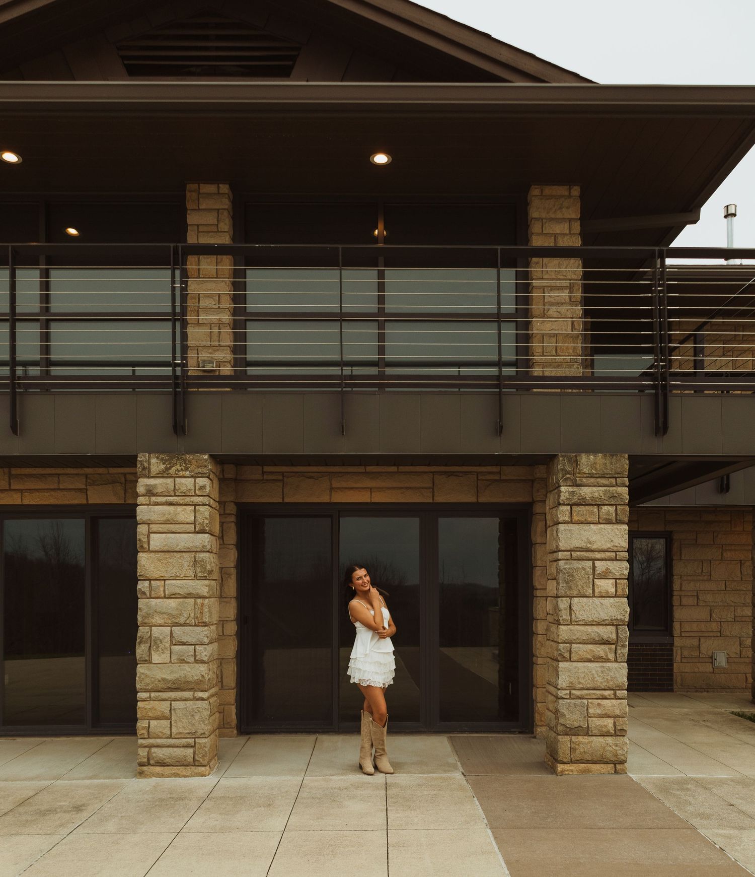 Woman in white dress posing in front of a modern stone building with dark glass doors and upper balcony.