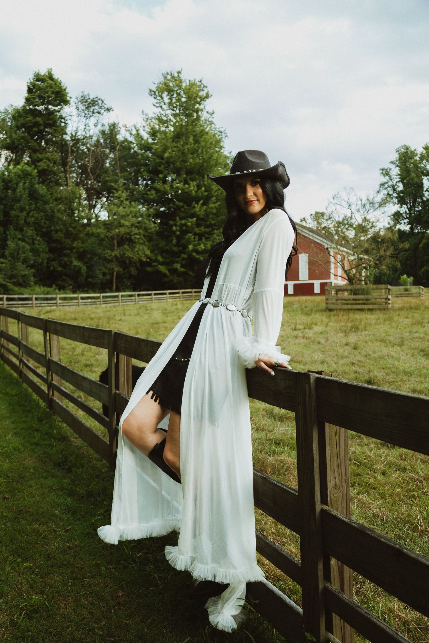 A flowing white dress billows in the wind as someone climbs over a rustic wooden fence in a pastoral field.