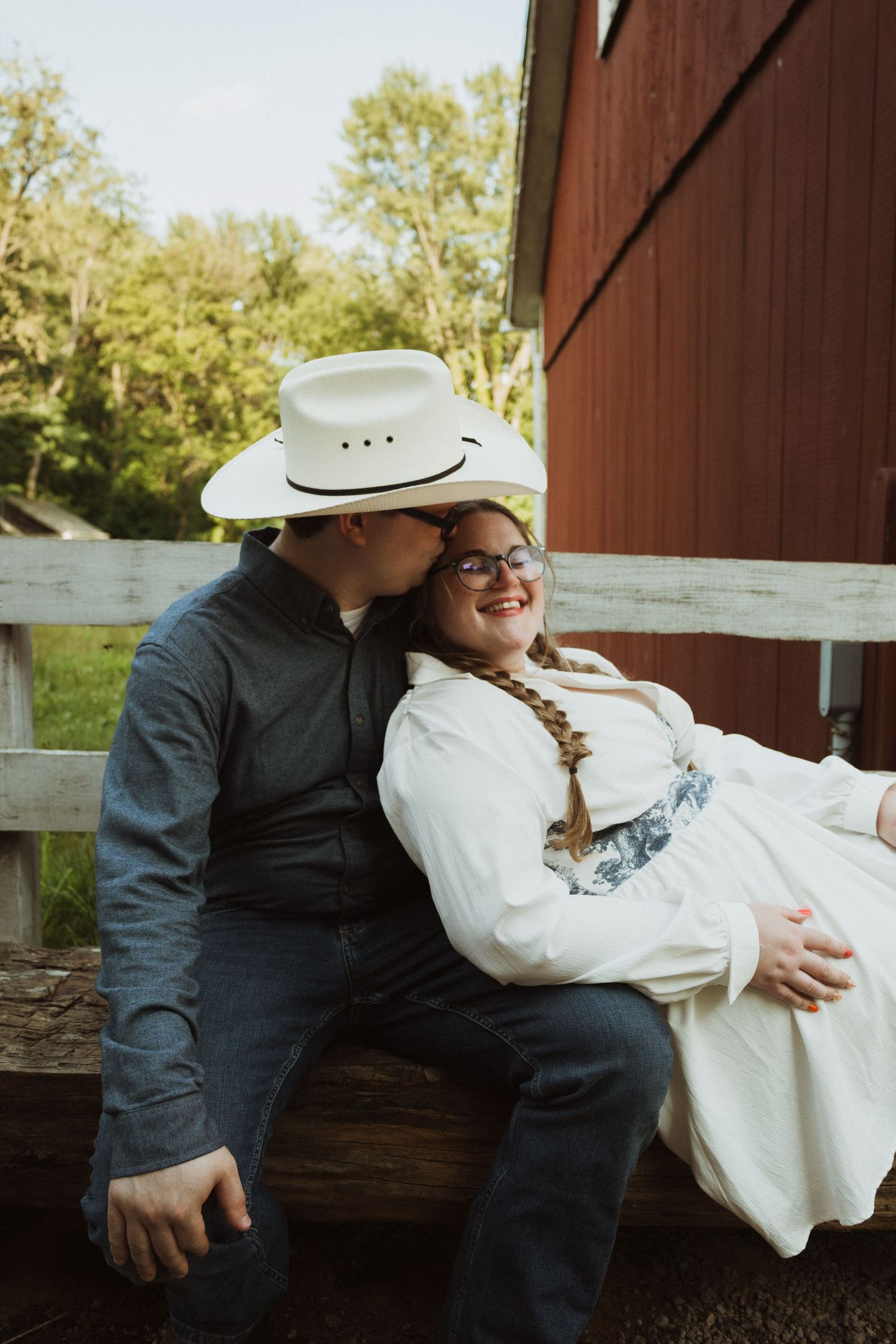 A couple in rustic western attire shares tender moments while sitting against a red barn wall.