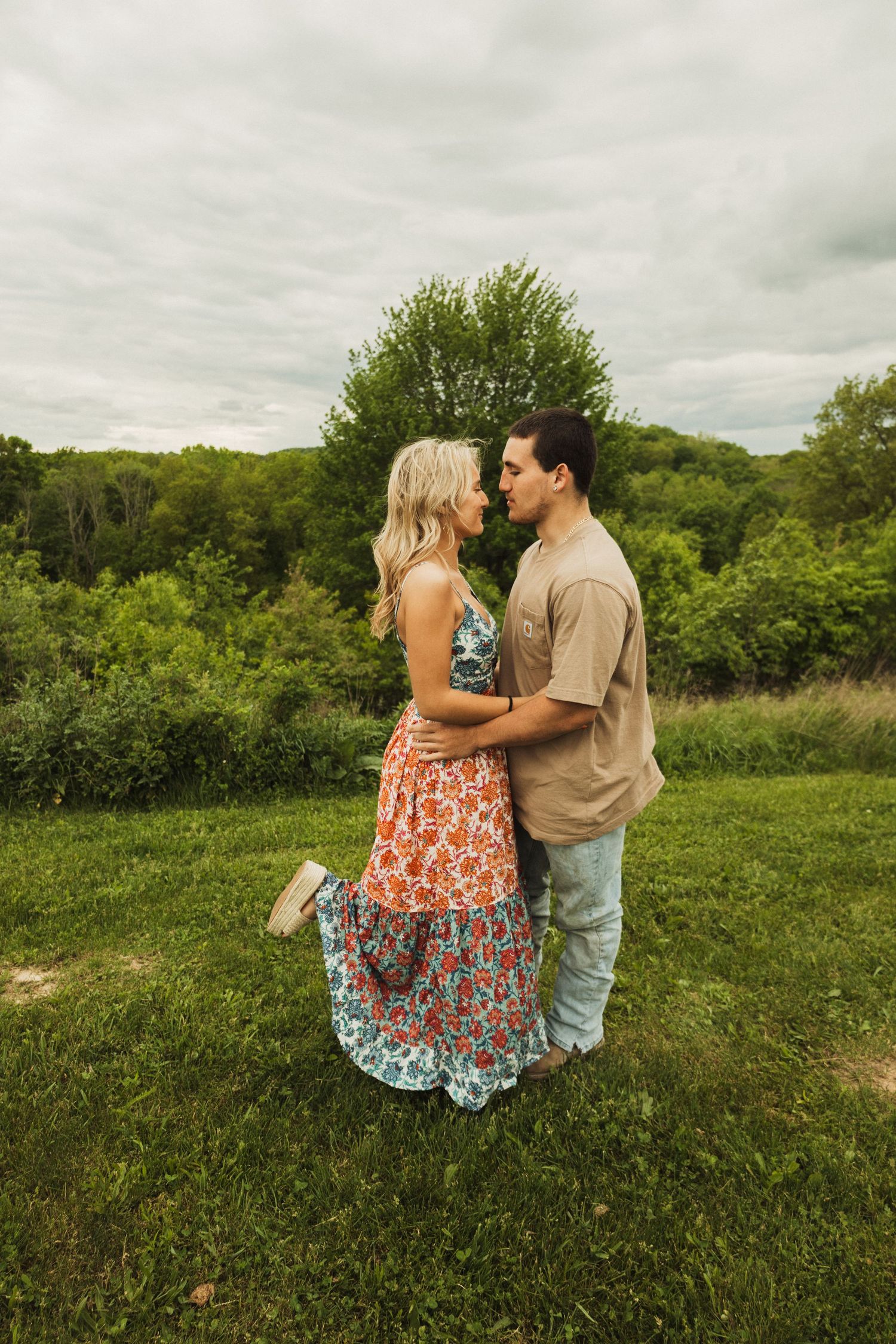 A couple shares an intimate moment while standing in a meadow with green trees in the background.