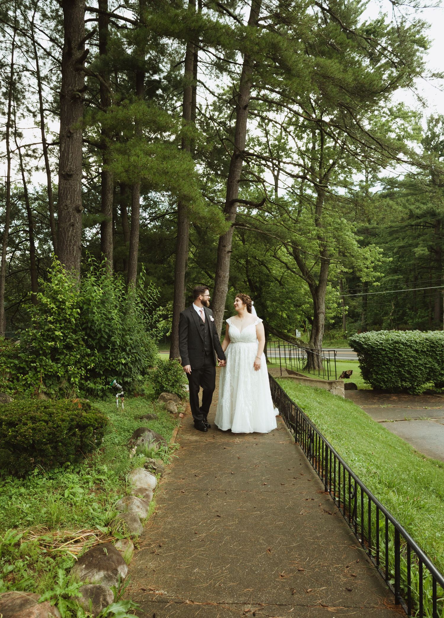 A couple walks along a winding garden path surrounded by lush greenery and tall pine trees.