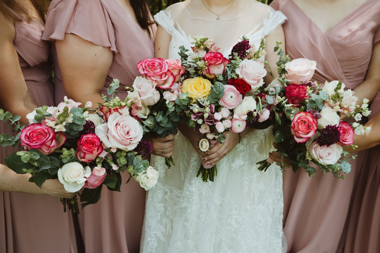 A row of pink and white floral wedding bouquets with roses and mixed flowers held against mauve bridesmaid dresses.
