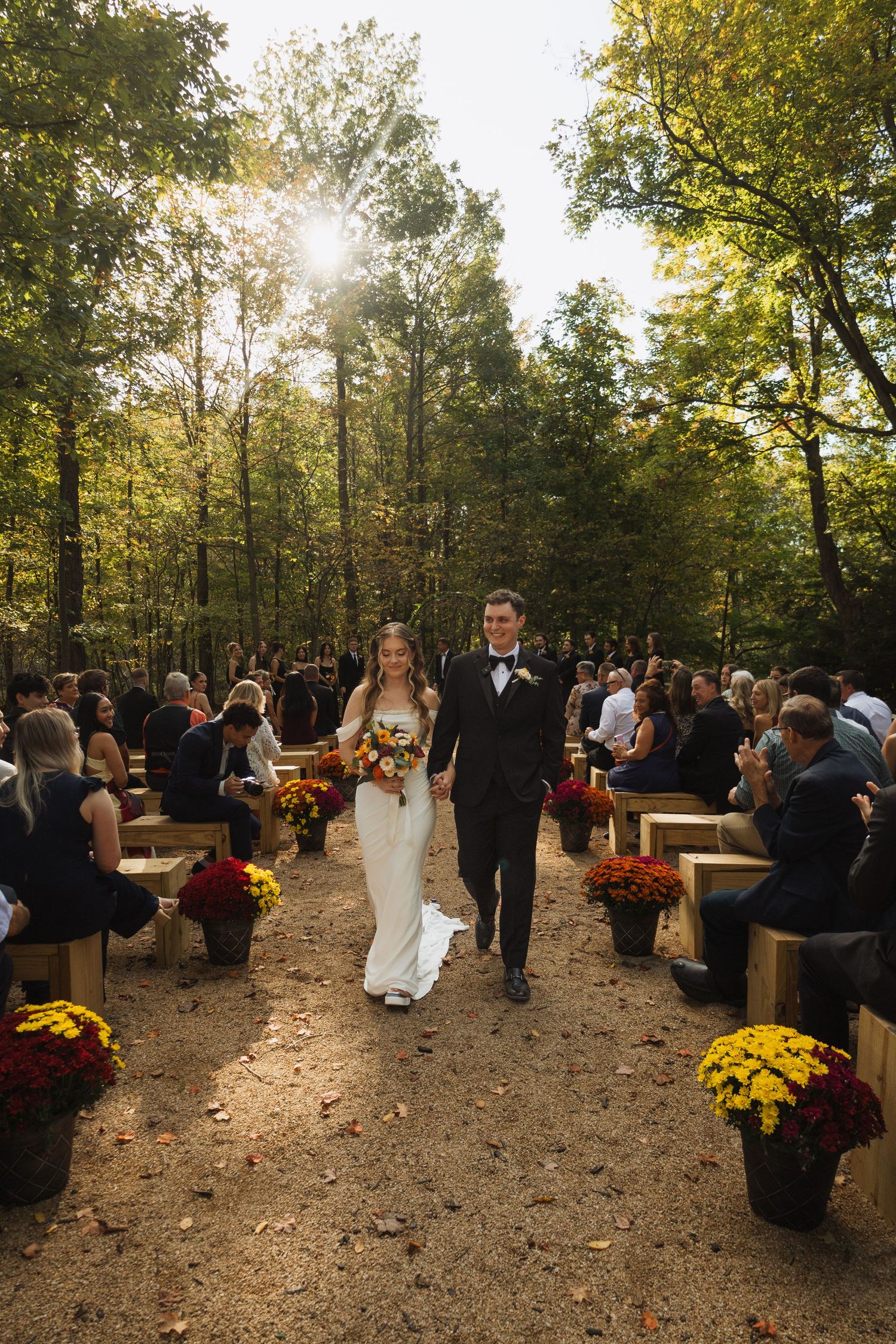Guests seated on wooden benches at an autumn outdoor wedding ceremony surrounded by trees and mums.