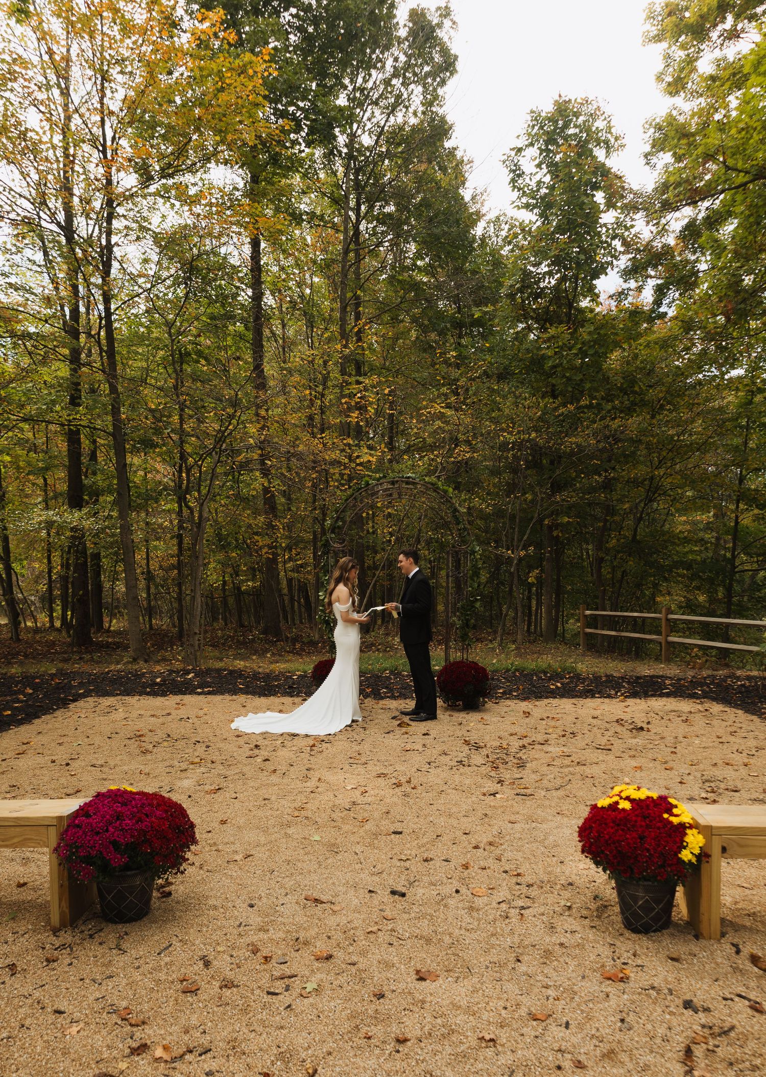 Fall wedding ceremony setup with red floral arrangements on a sandy clearing surrounded by autumn trees.