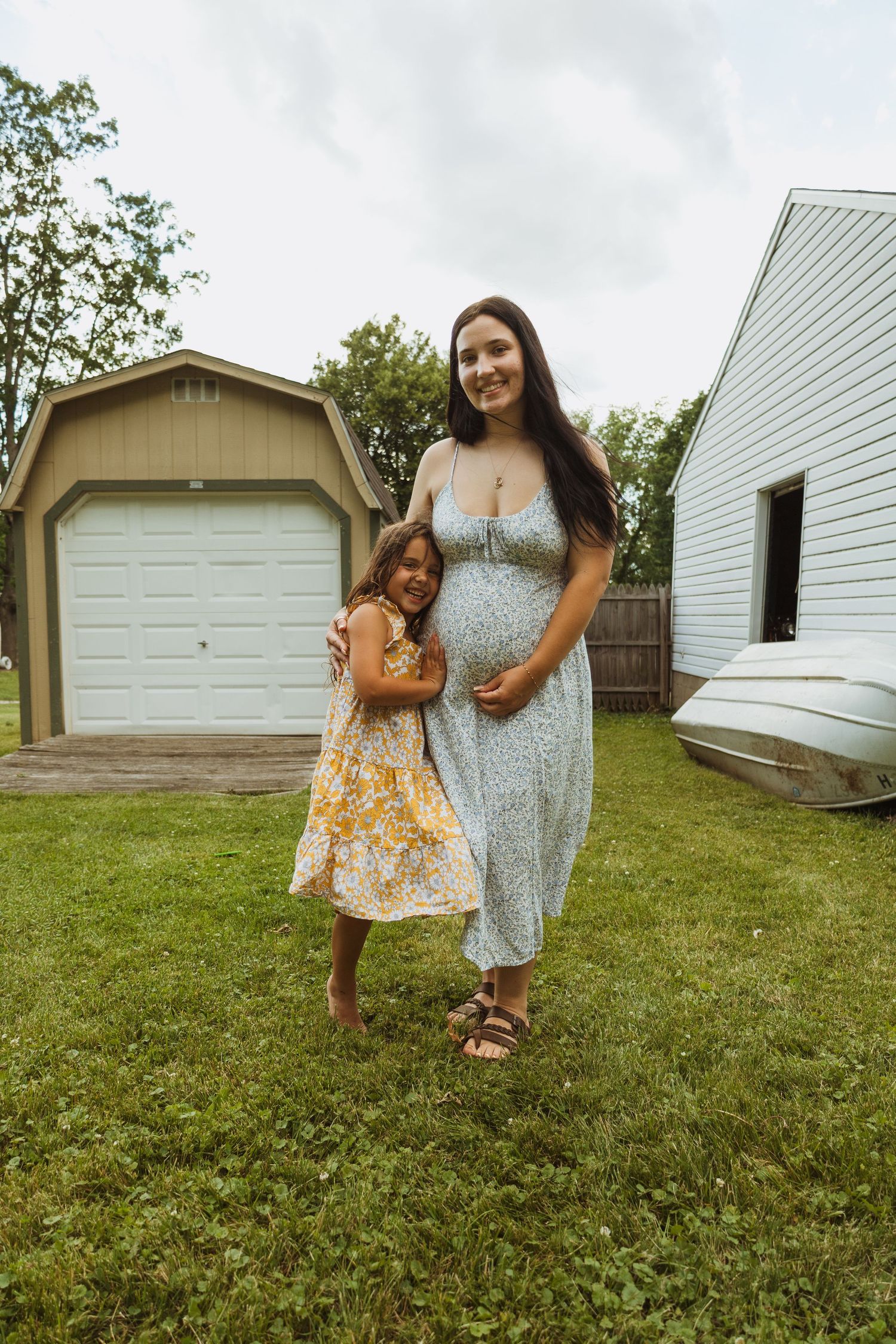 Pregnant woman in floral dress hugging young girl in yellow dress, standing on green lawn near garage.
