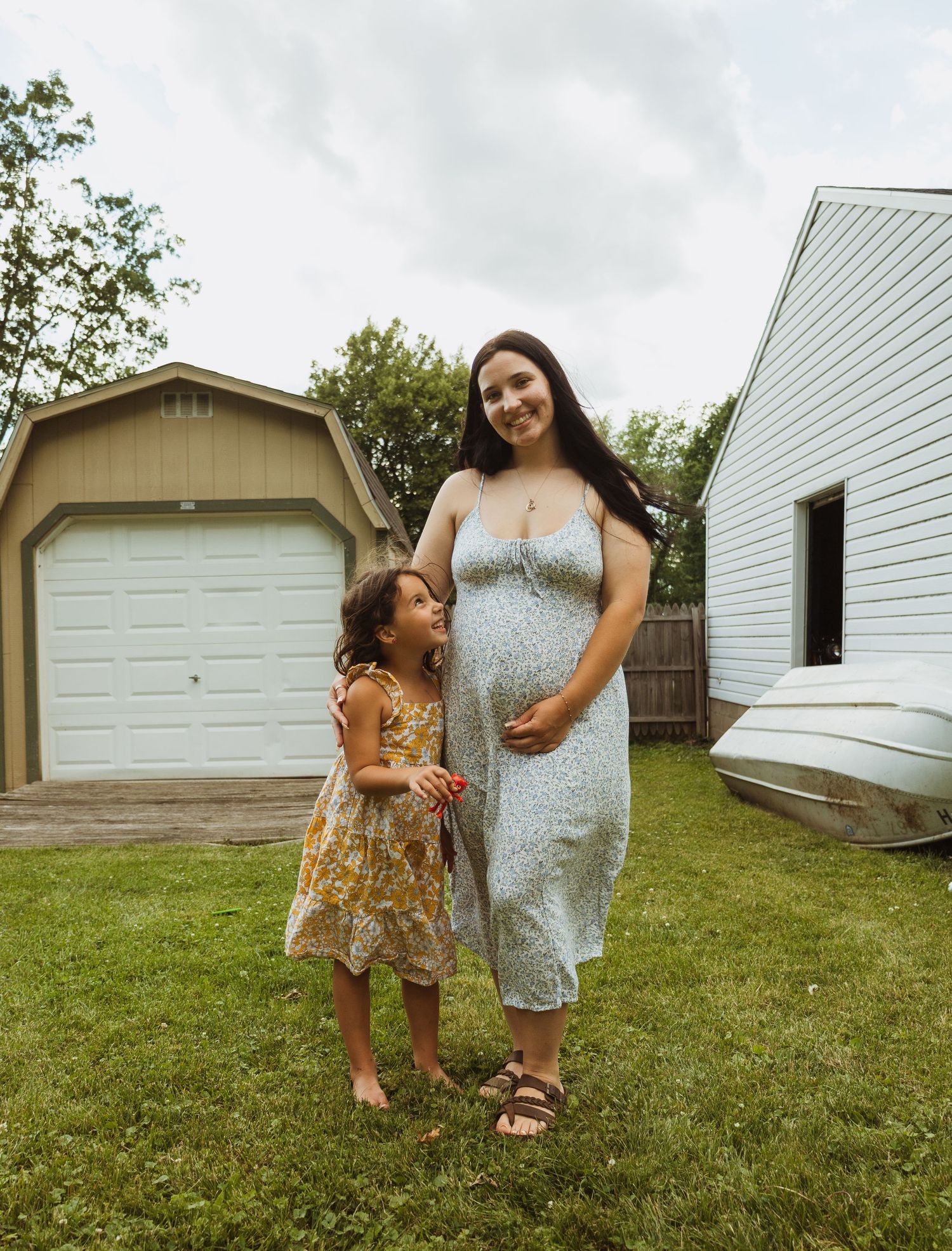 Pregnant woman in floral dress stands smiling with young girl in yellow dress on green lawn near garage.