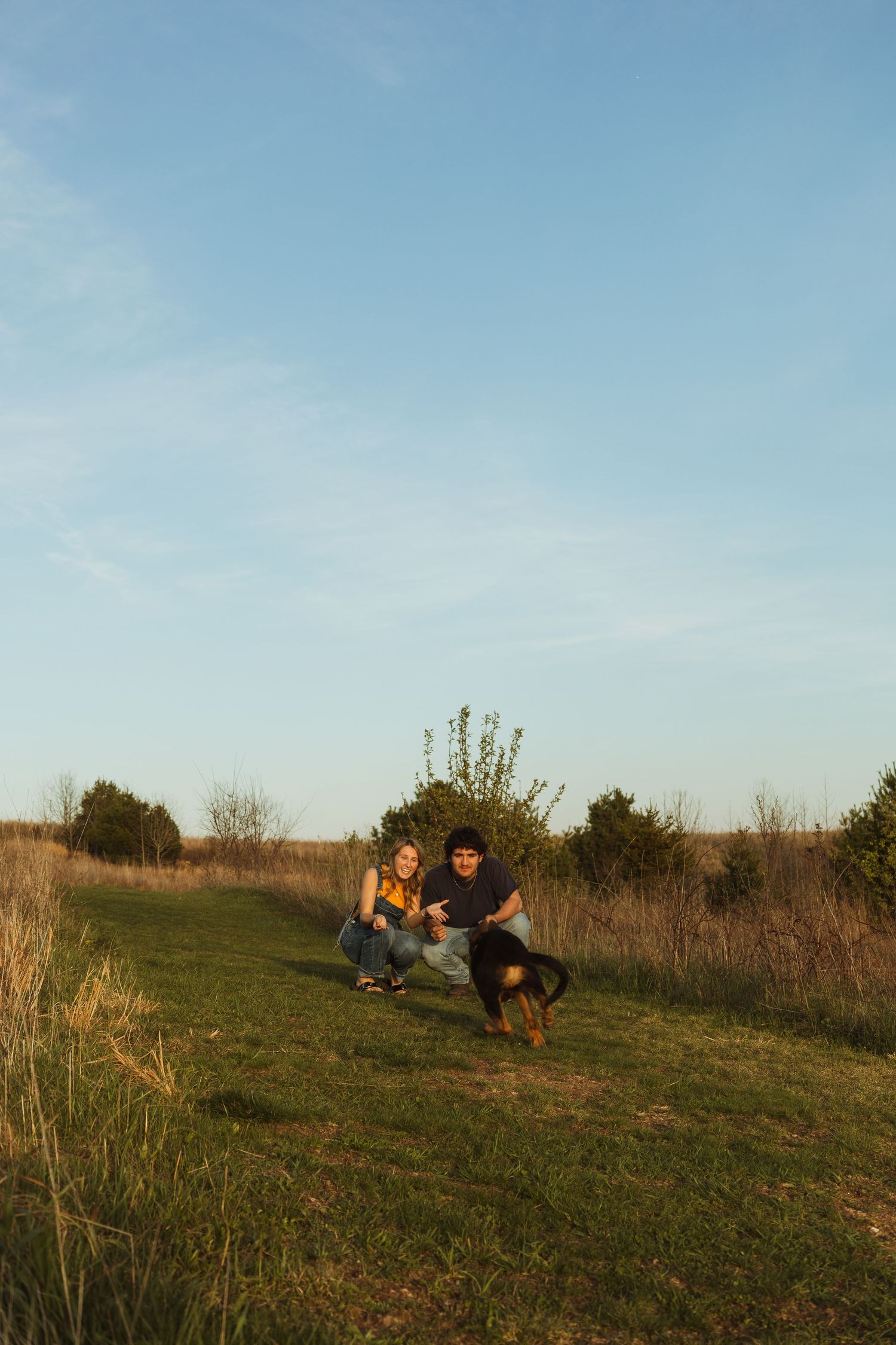 Couple crouching with their dog on green grass field at golden hour with blue sky and trees in background.